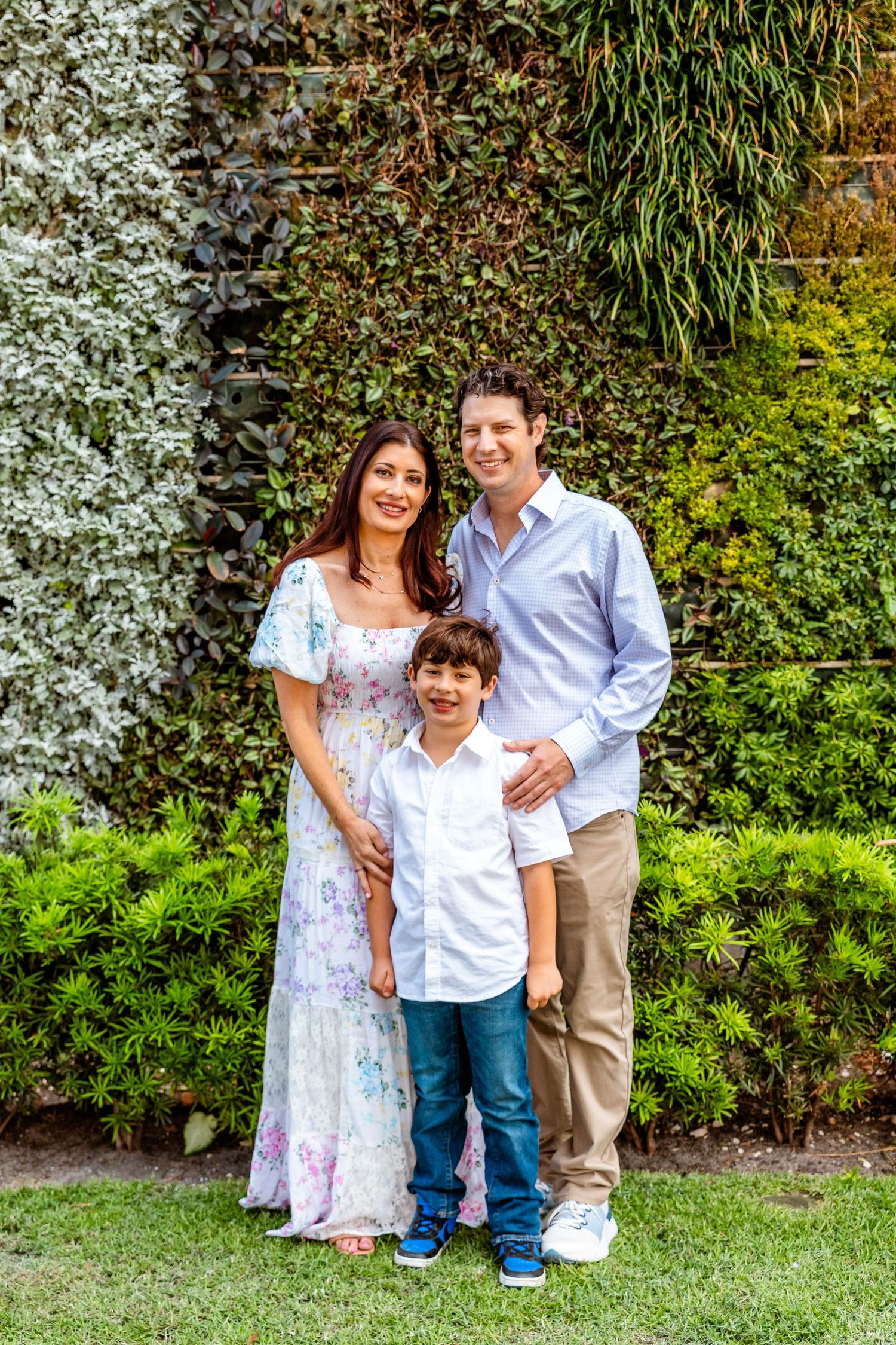 Smiling family of three posing outdoors in front of a lush green ivy-covered garden wall.
