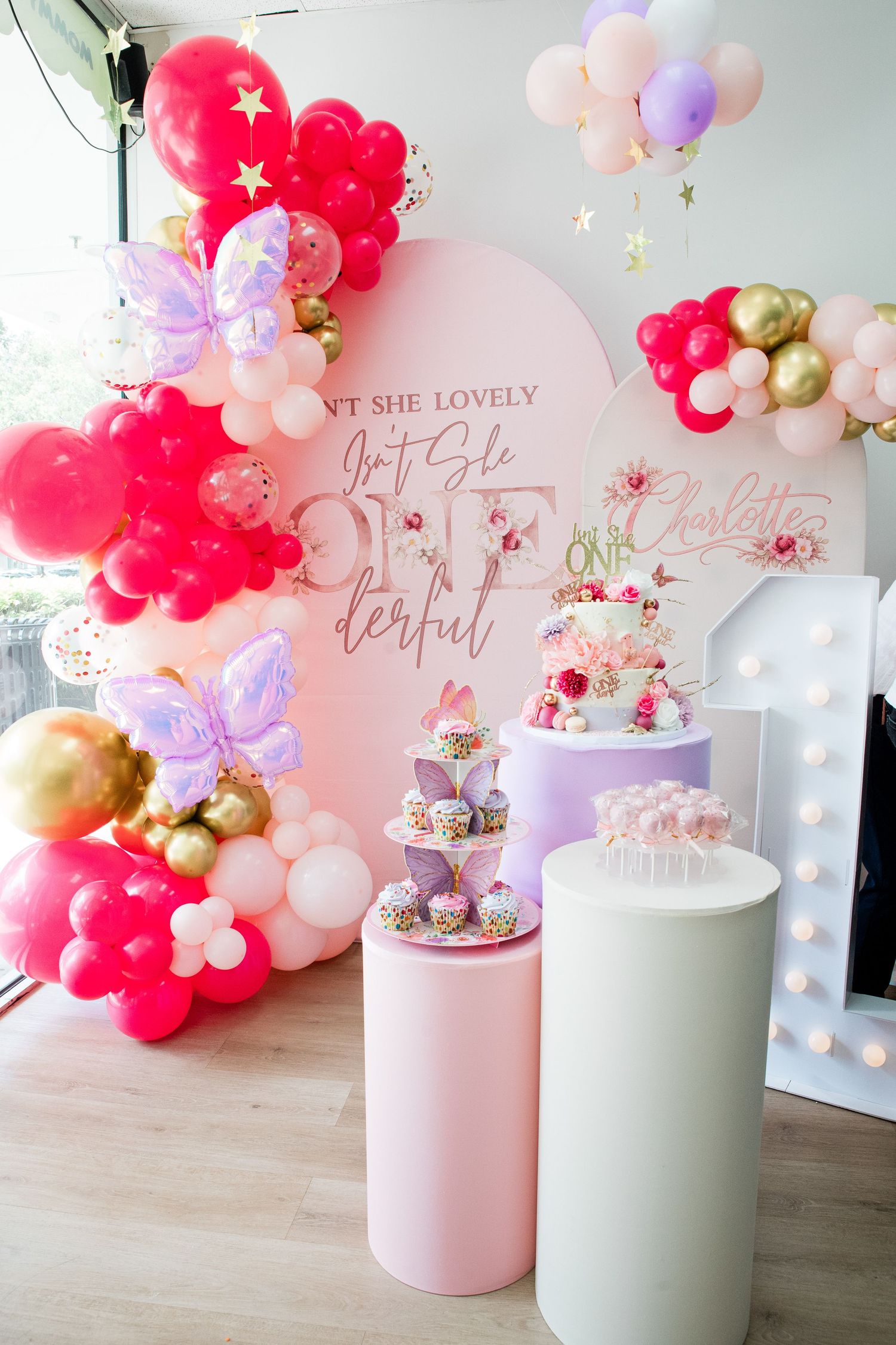Elegant first birthday party setup with pink balloon arch, butterfly decorations, dessert tower, and marquee number one display.