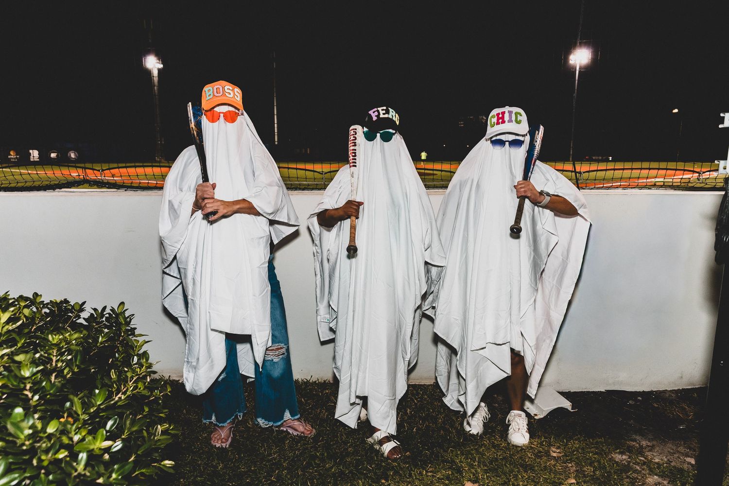 Three people dressed in white ghost costumes with masks pose together at night against a white wall.