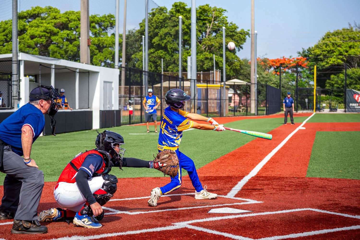 Young baseball batter in blue and gold uniform swinging at pitch during youth little league game on red turf field.
