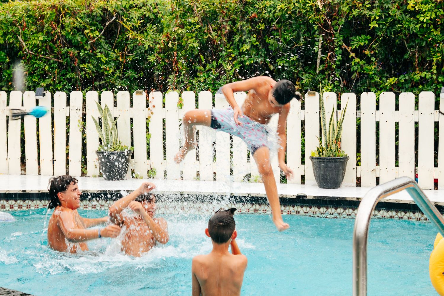 People playing and jumping into a backyard swimming pool surrounded by a white picket fence on a sunny day.