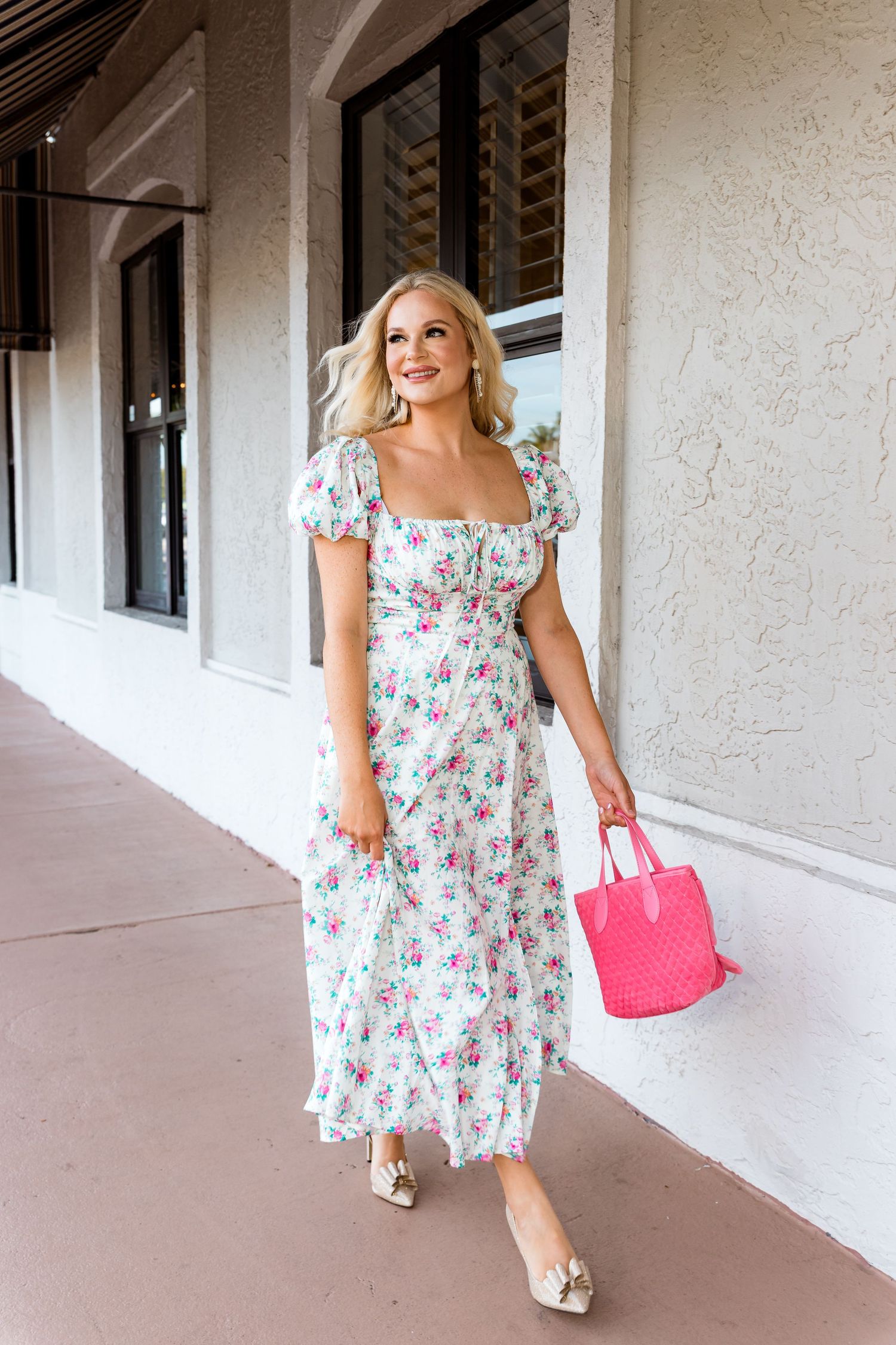 Woman in floral midi dress walking with pink handbag along covered walkway.