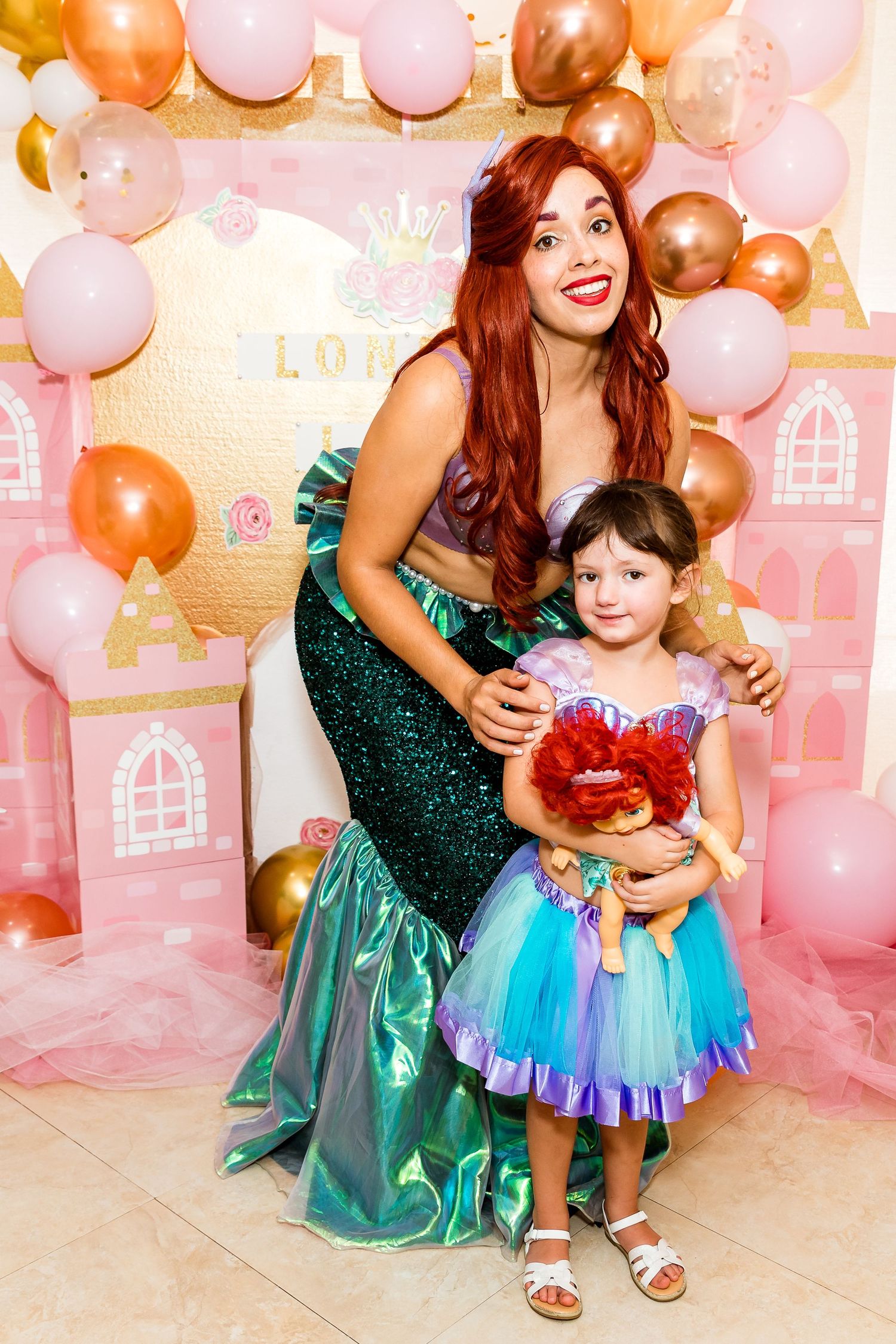Princess performer in sparkly teal gown poses with child at magical party backdrop with pink and gold balloons.