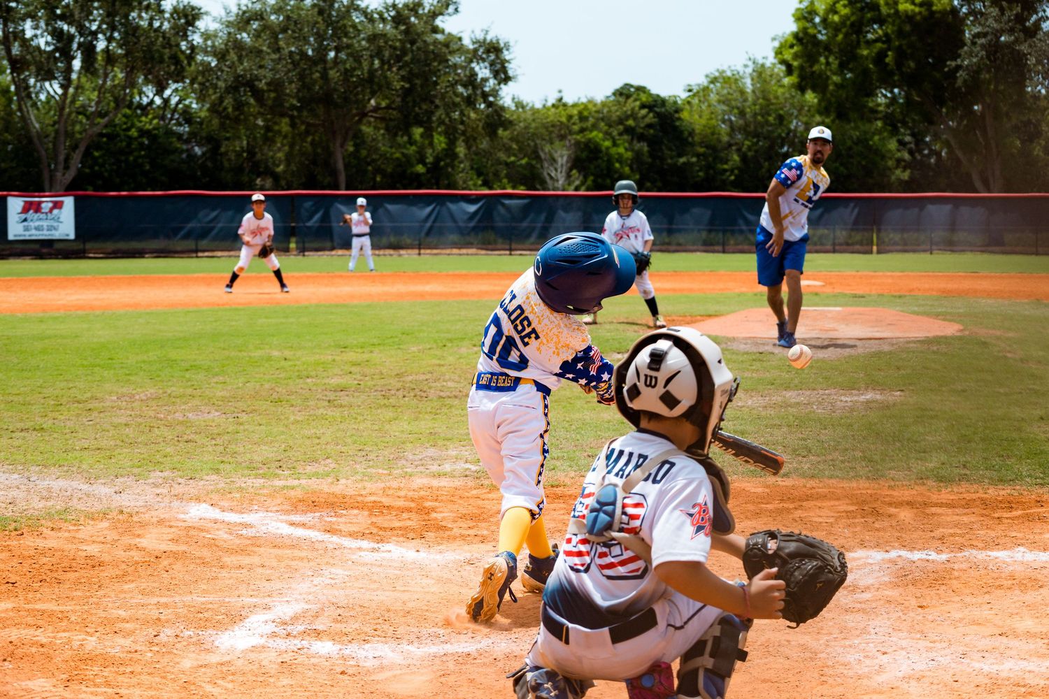 Youth baseball game in progress with batter at home plate and catcher crouched behind as pitcher delivers the ball.
