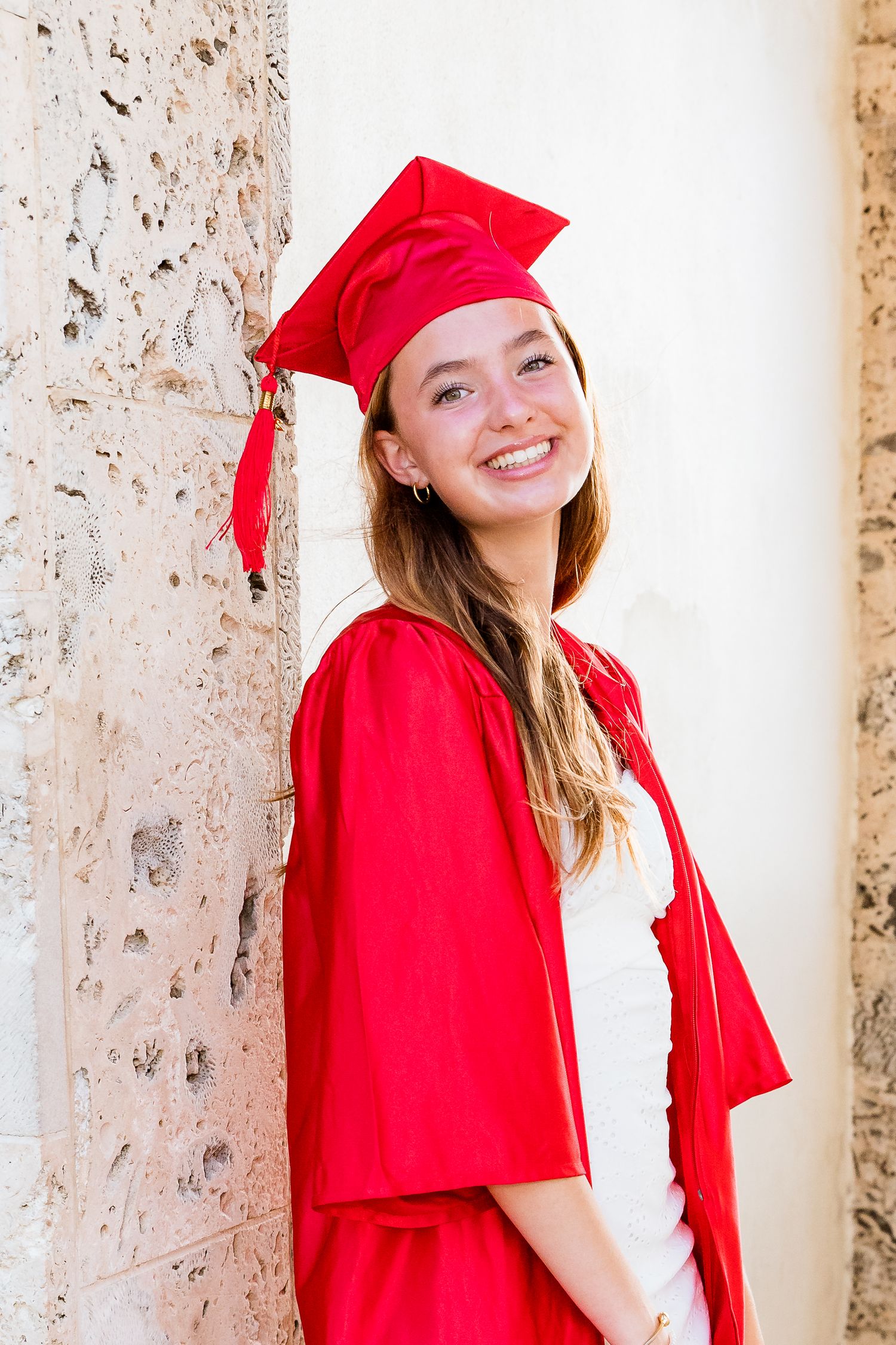 Graduate in bright red cap and gown smiles joyfully while posing against a textured stone wall.