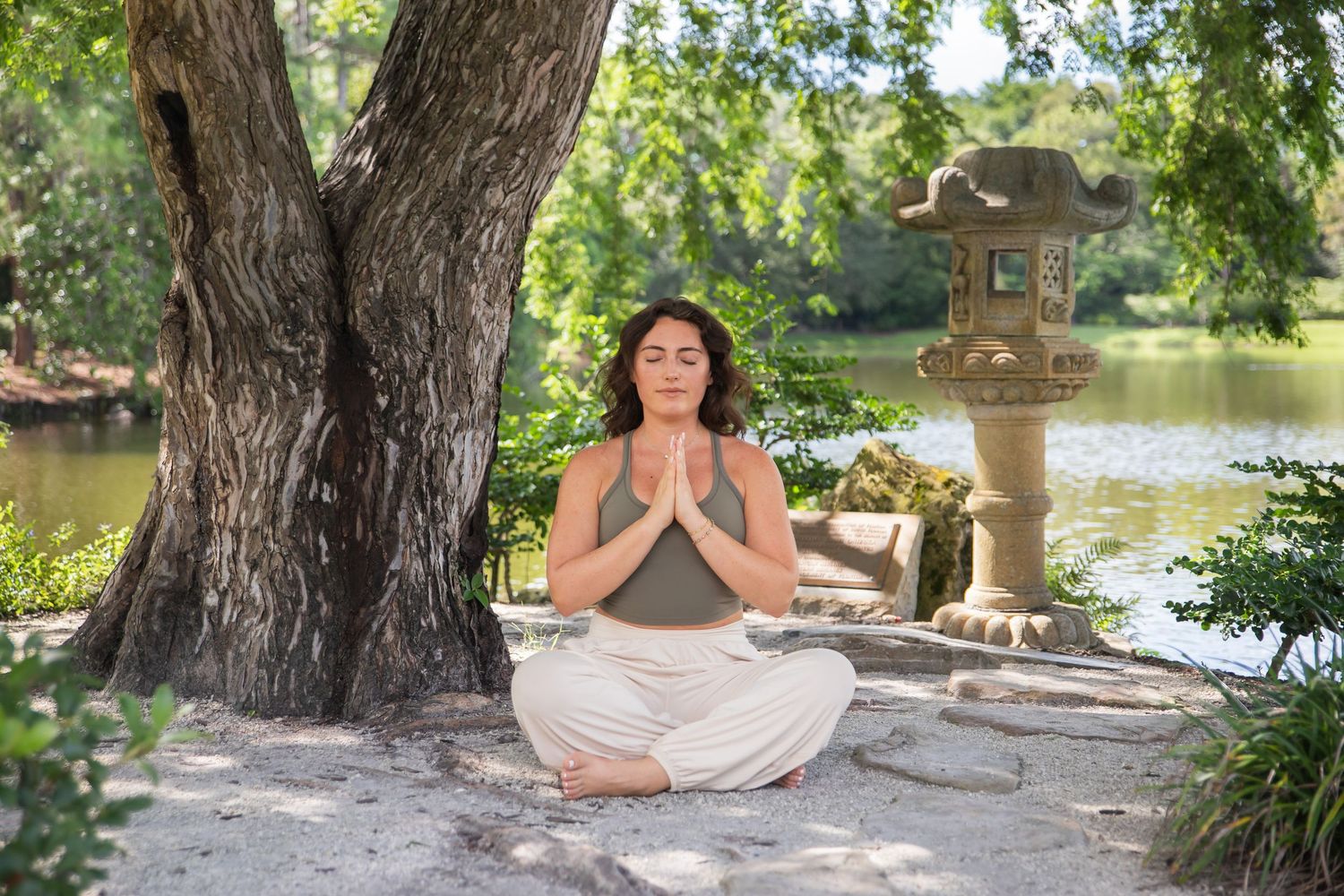 A person sits in lotus position practicing meditation near a lake surrounded by trees and nature scenery.