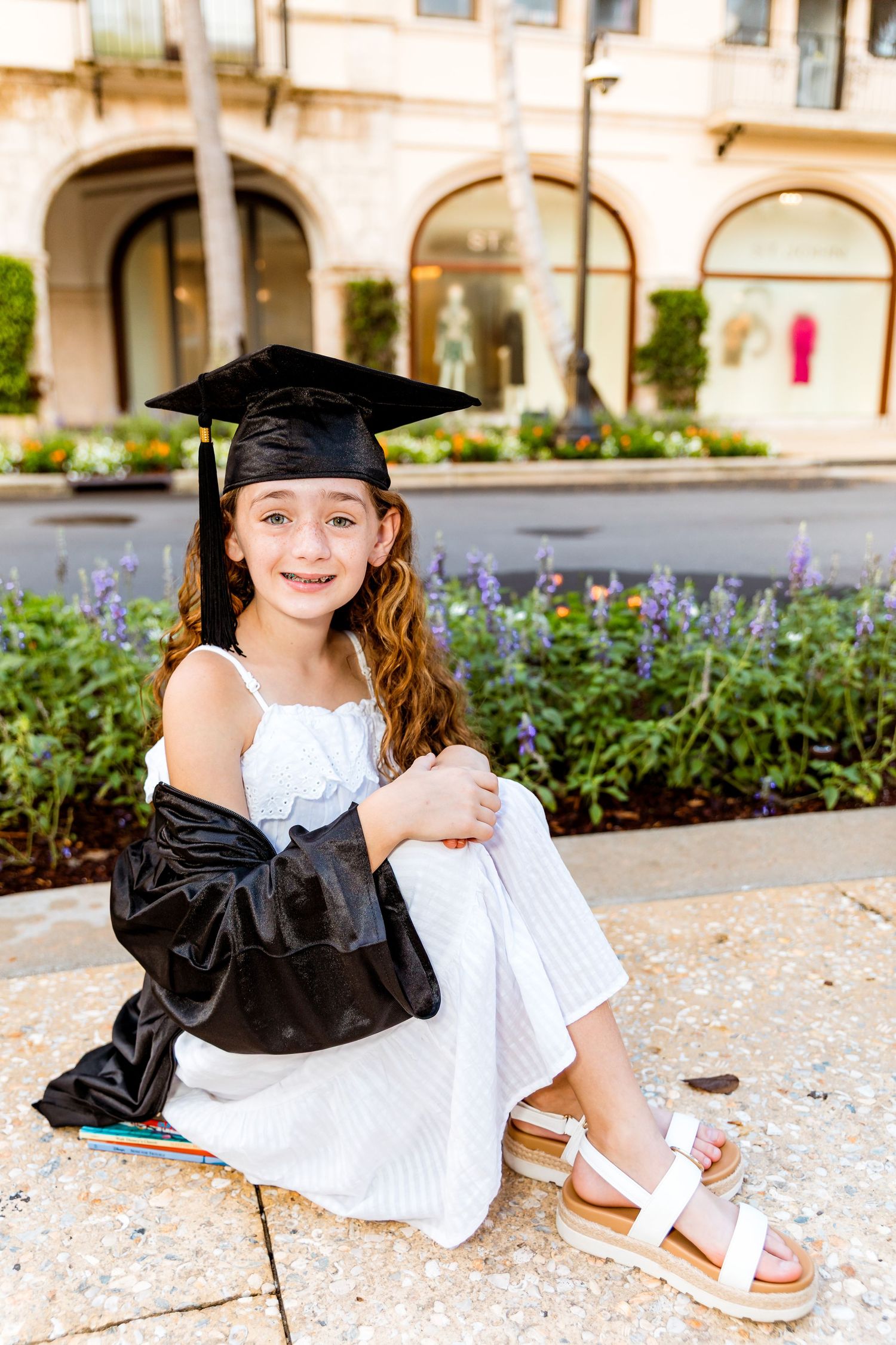 Young girl in graduation cap and gown sitting outdoors in white dress, celebrating academic milestone.