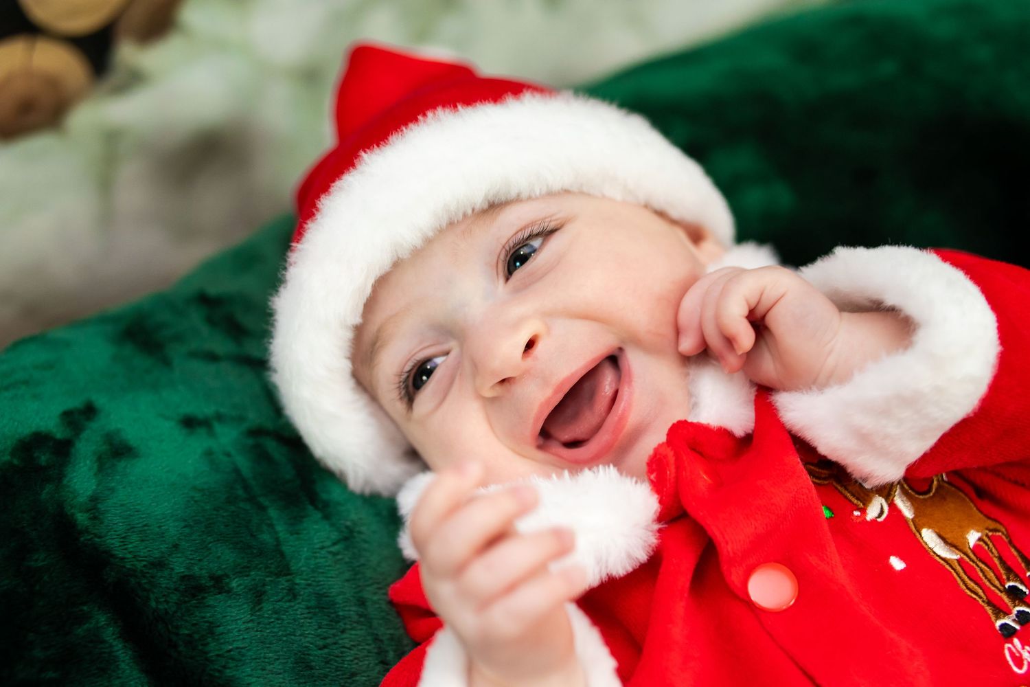 Baby in red Santa outfit and hat smiles joyfully while lying on green velvet blanket.