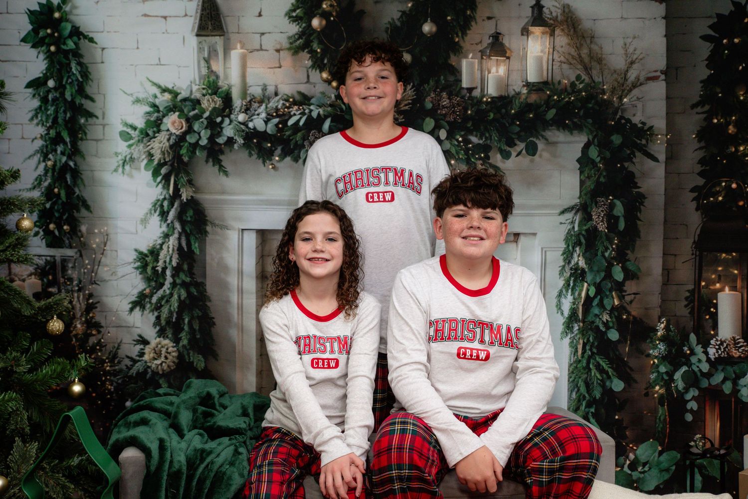 Two people in matching white Christmas sweaters and plaid pajama pants pose on a white bench with holiday decor.