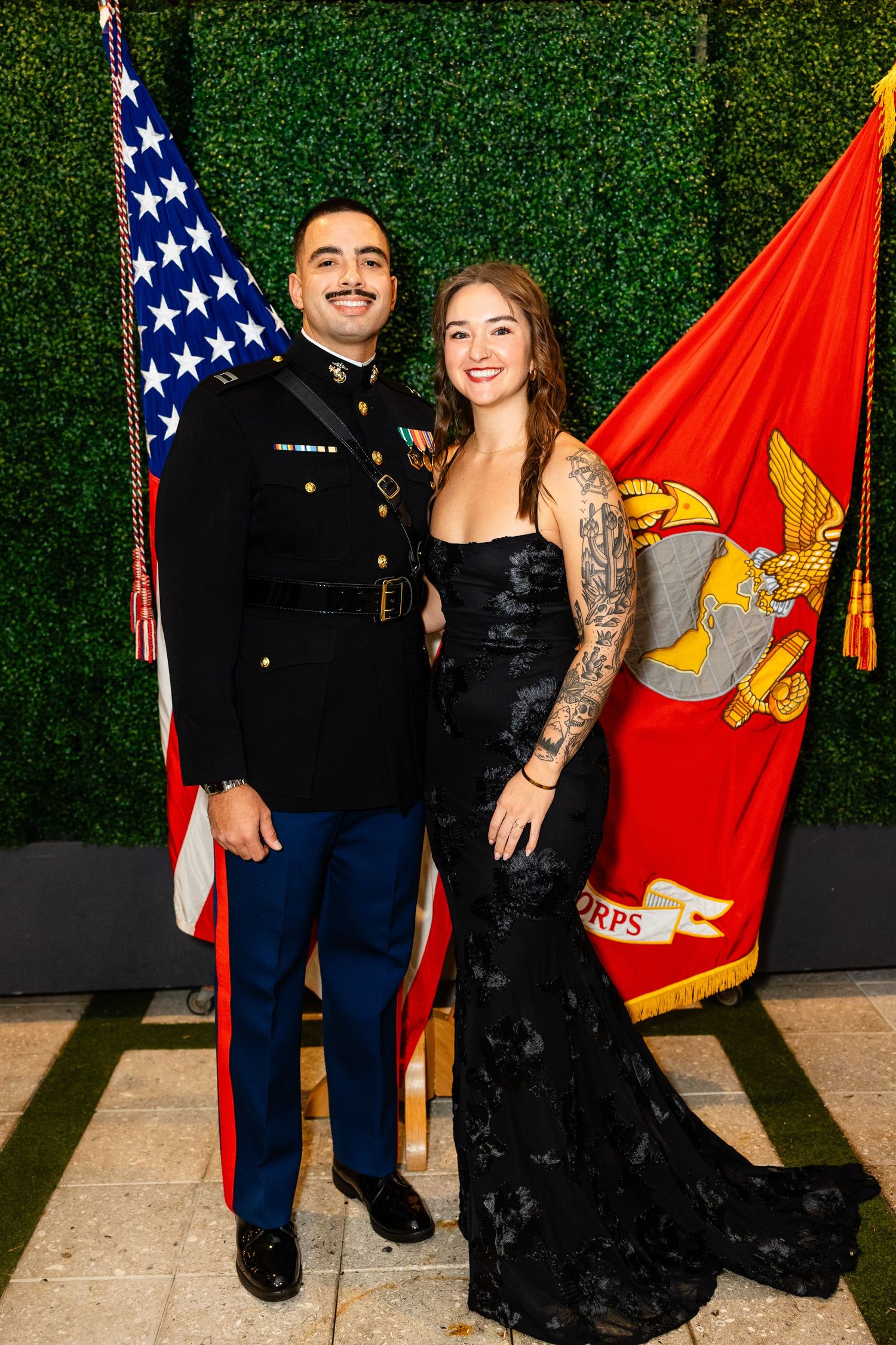 A couple poses in front of a green hedge backdrop with American and Marine Corps flags at a formal military event.