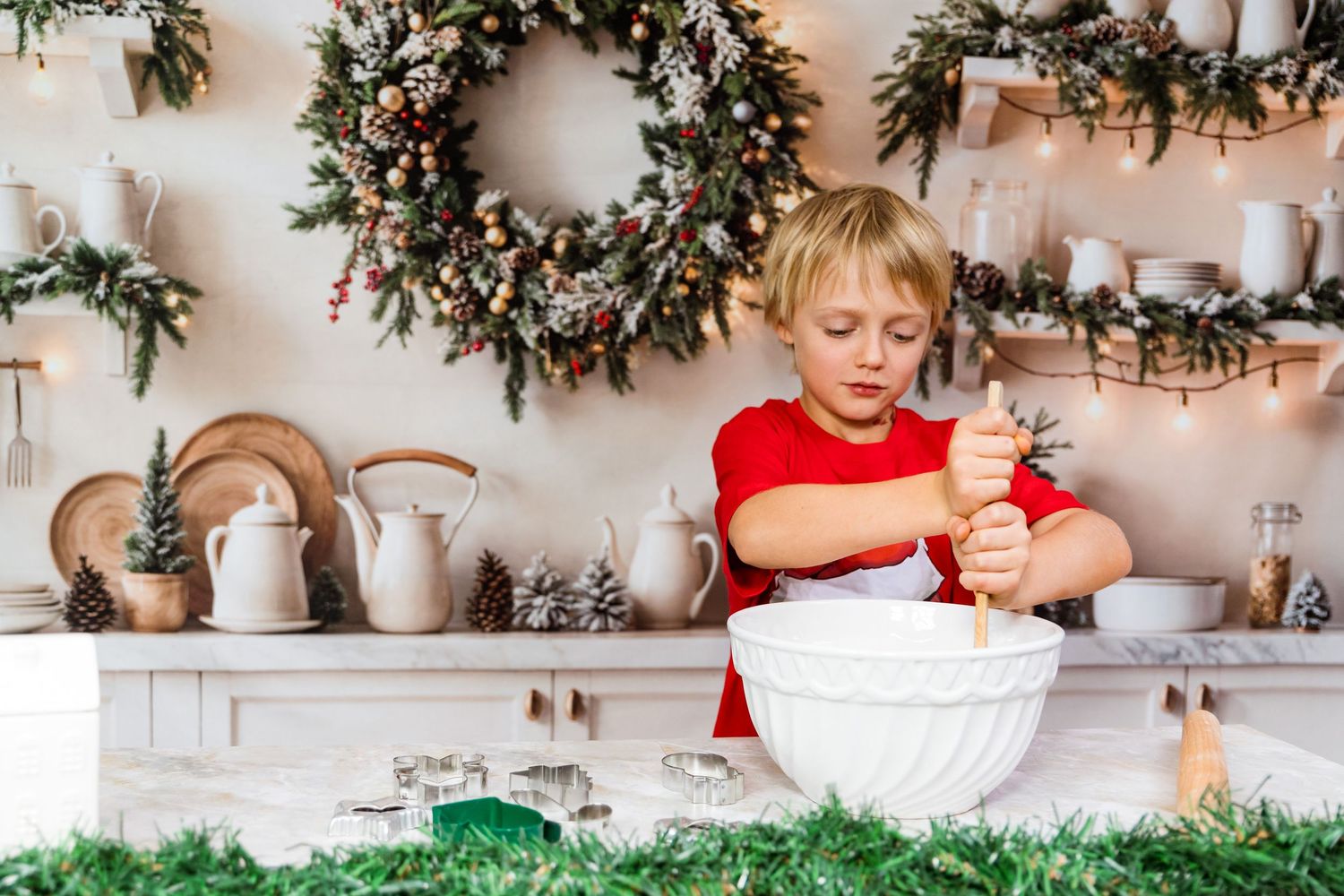 Child in red shirt baking Christmas cookies in kitchen with festive wreath and decorations on wall.