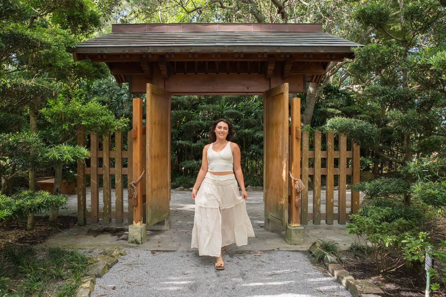 Someone in a white dress walks through a wooden garden gate surrounded by lush greenery in a Japanese-inspired garden.