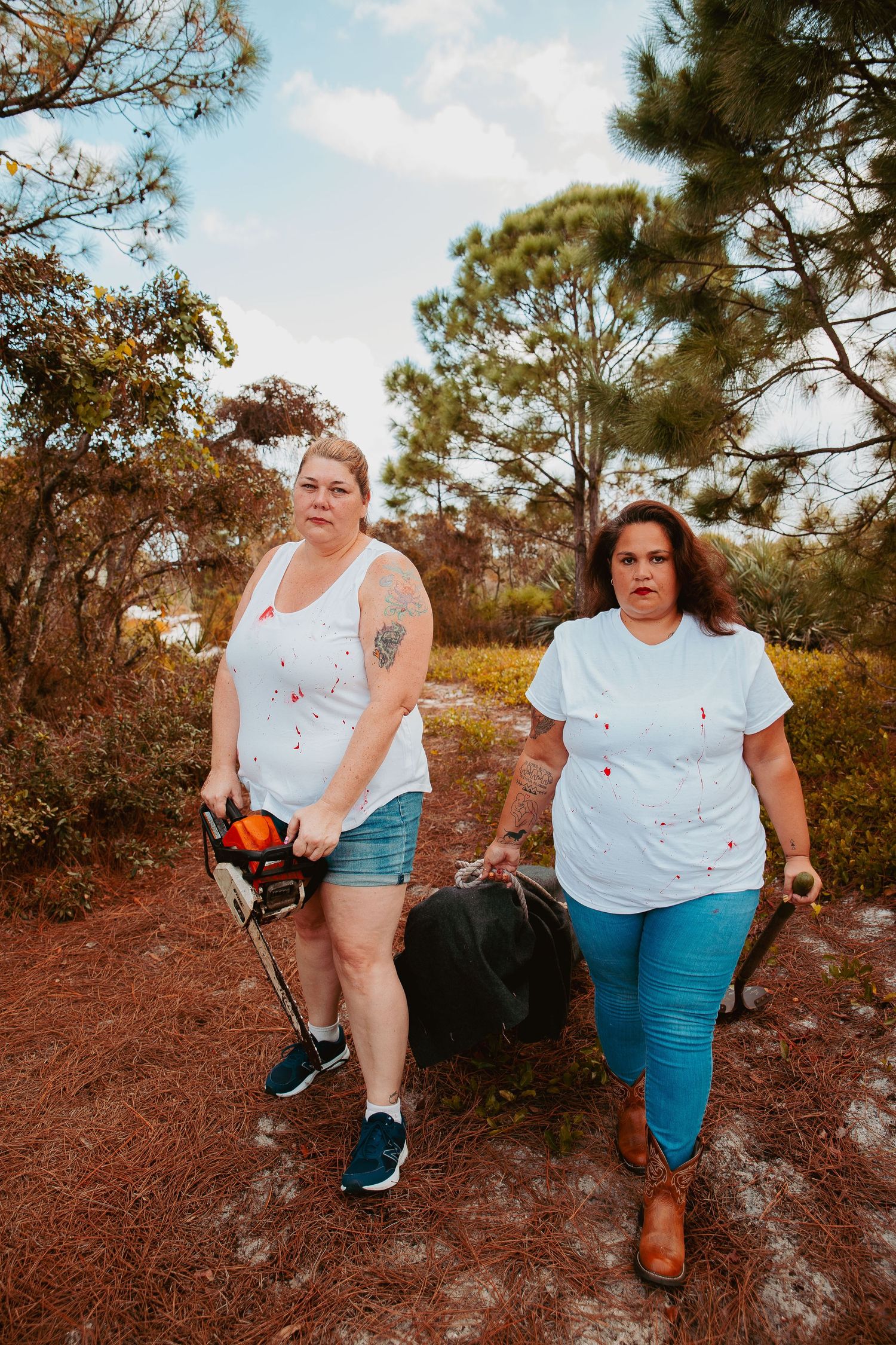 Two people walking on a dirt path carrying gardening tools through a wooded area with pine trees.