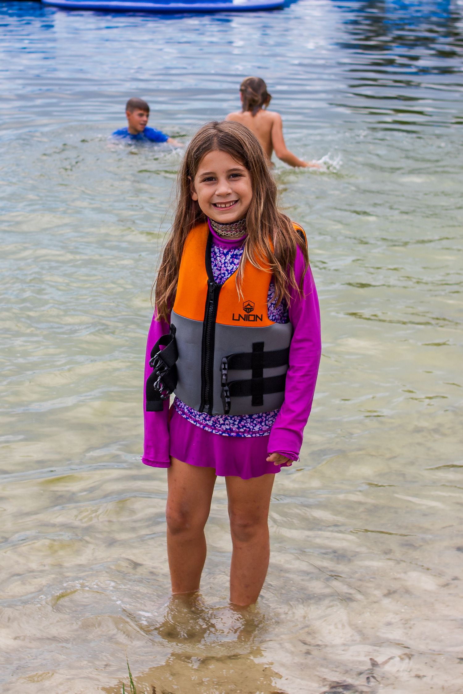 People wearing life vests standing in shallow clear water near the shore on a sunny day.