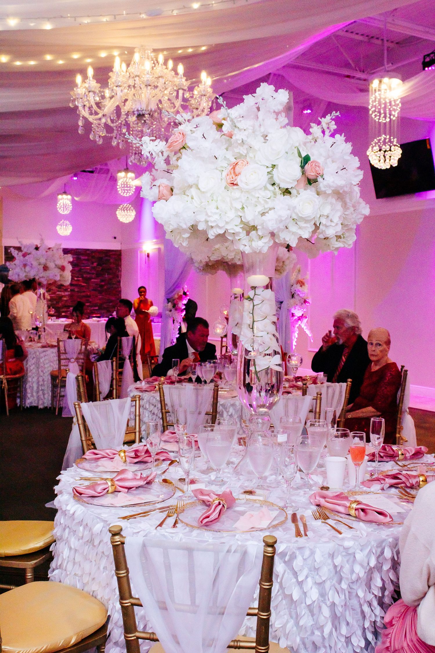 Luxurious wedding reception venue with crystal chandelier and tall white floral centerpieces.