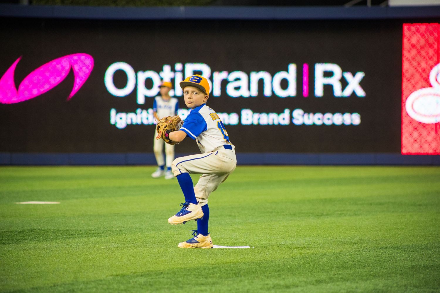 Baseball player demonstrates fielding position during practice session with OptiBrand Rx sign visible.