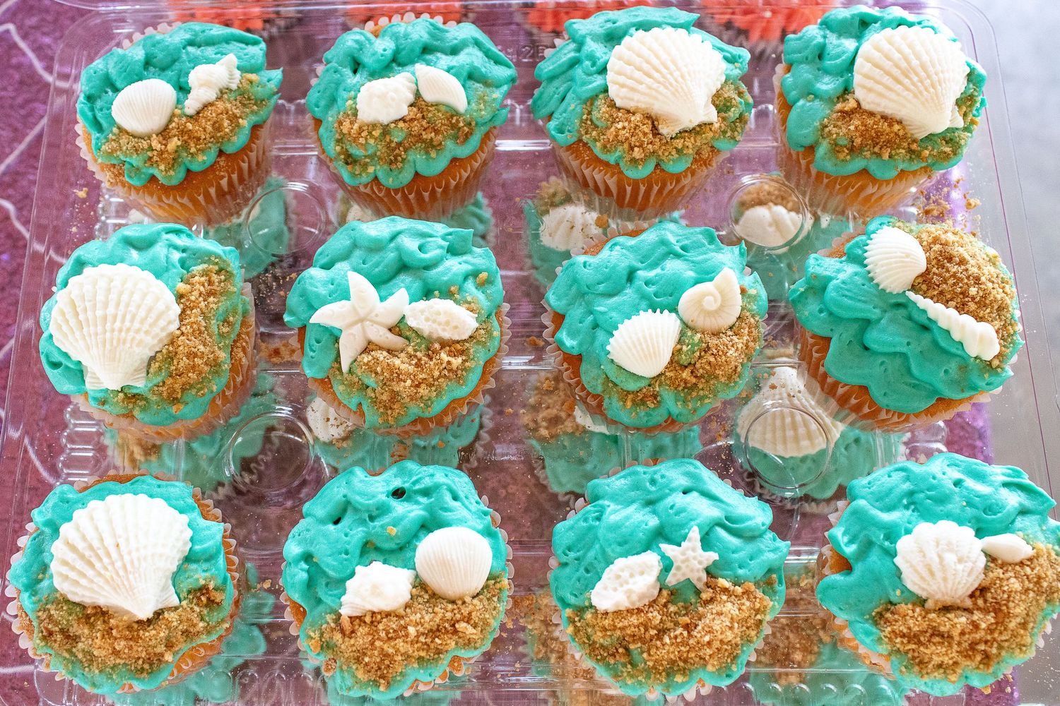 Ocean-themed cupcakes decorated with blue frosting, seashells, and edible sand on a display tray.