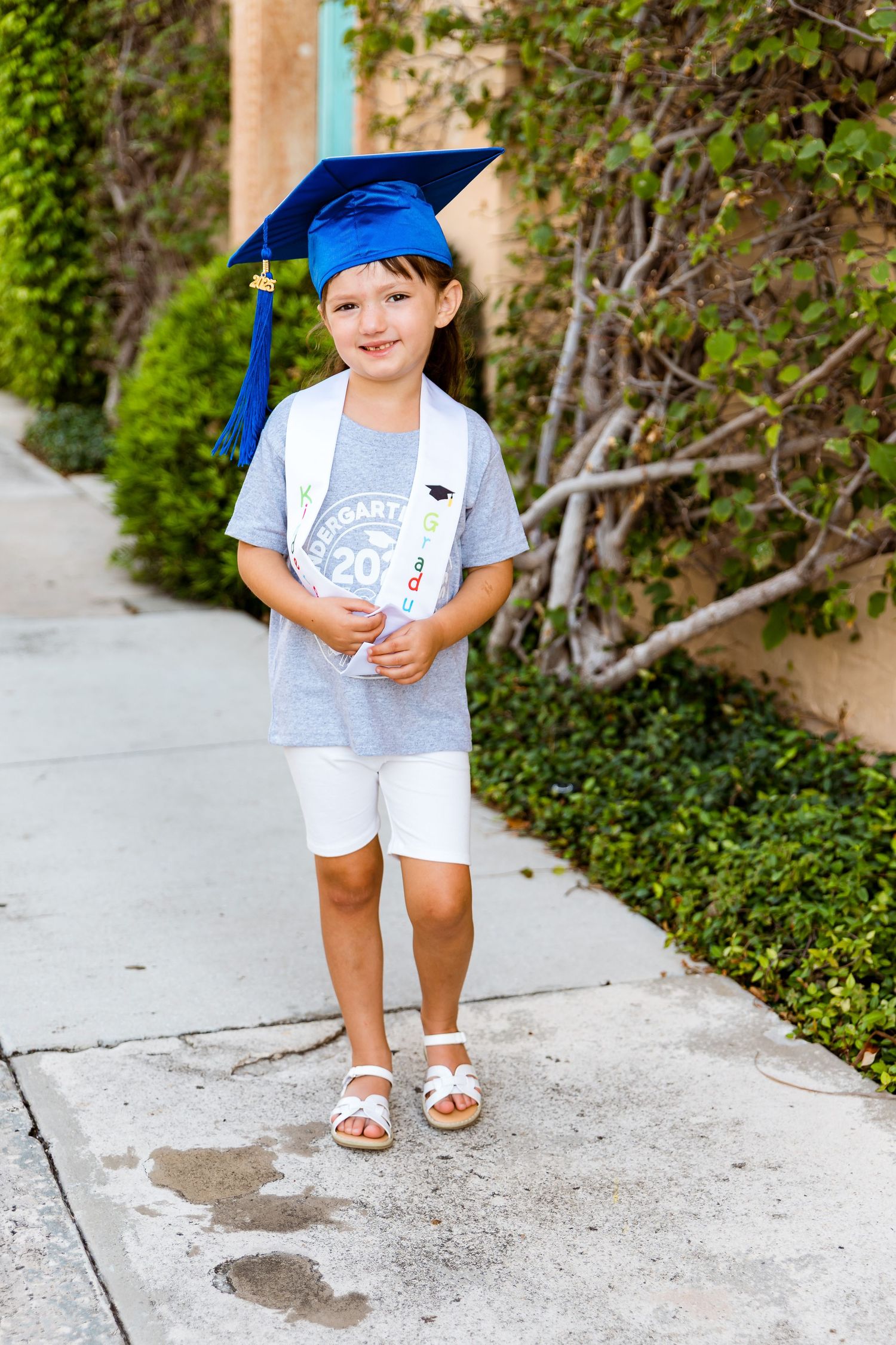 Young student wearing blue graduation cap, grey t-shirt and white shorts stands on sidewalk near greenery.