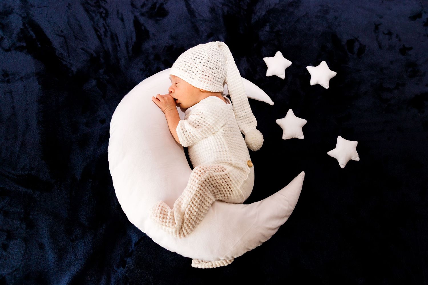 Newborn sleeping peacefully on white crescent moon cushion with decorative stars against navy backdrop.
