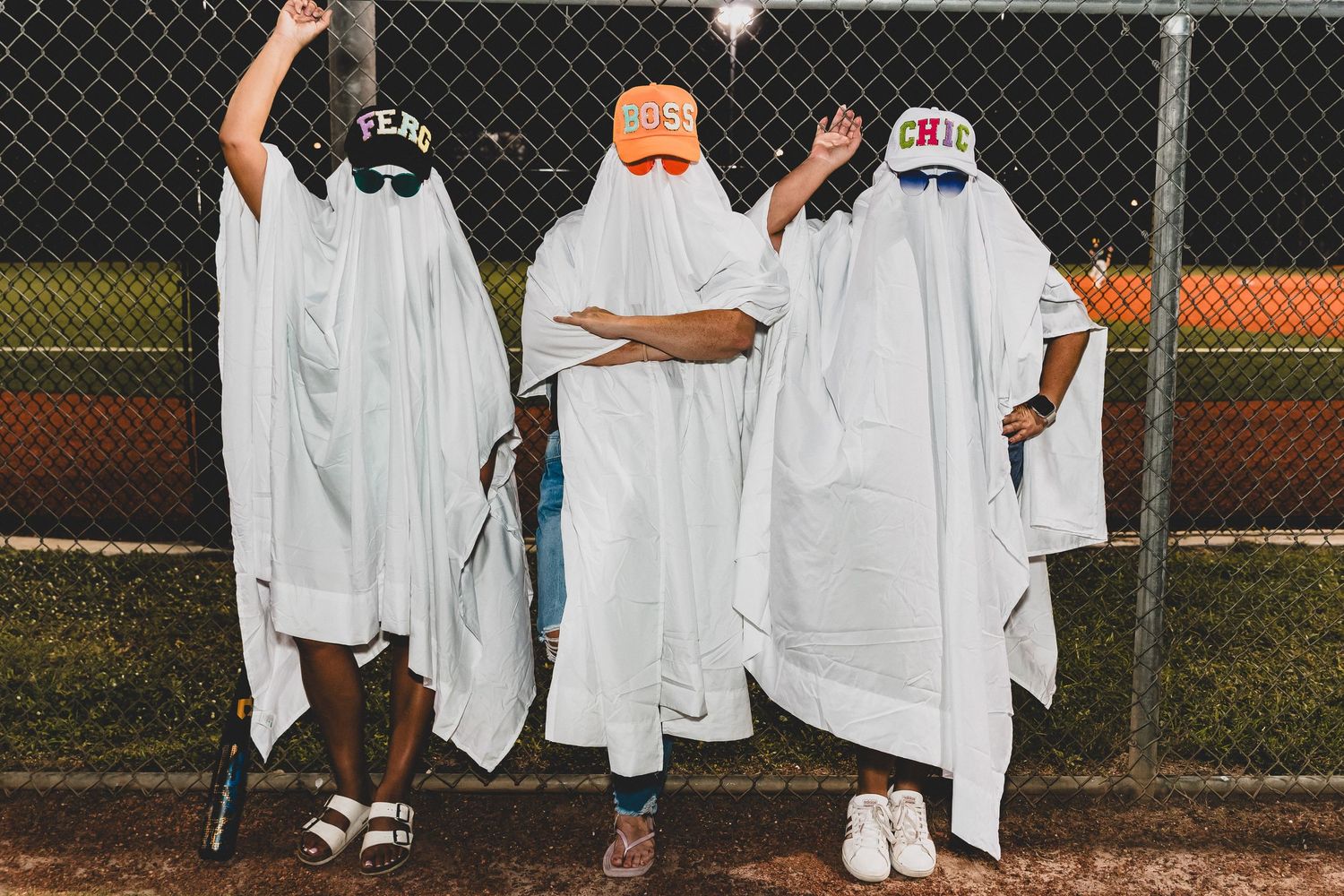 People dressed in white sheet costumes stand along chain link fence at night.