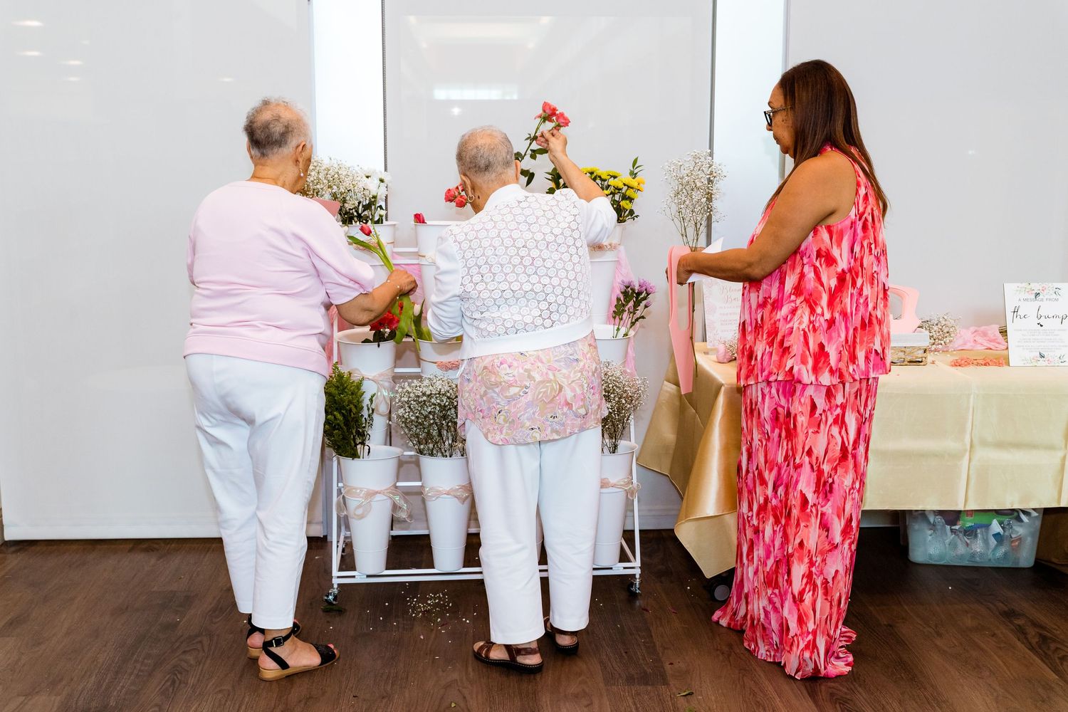 Women arranging floral decorations at an event with pink and white color scheme.