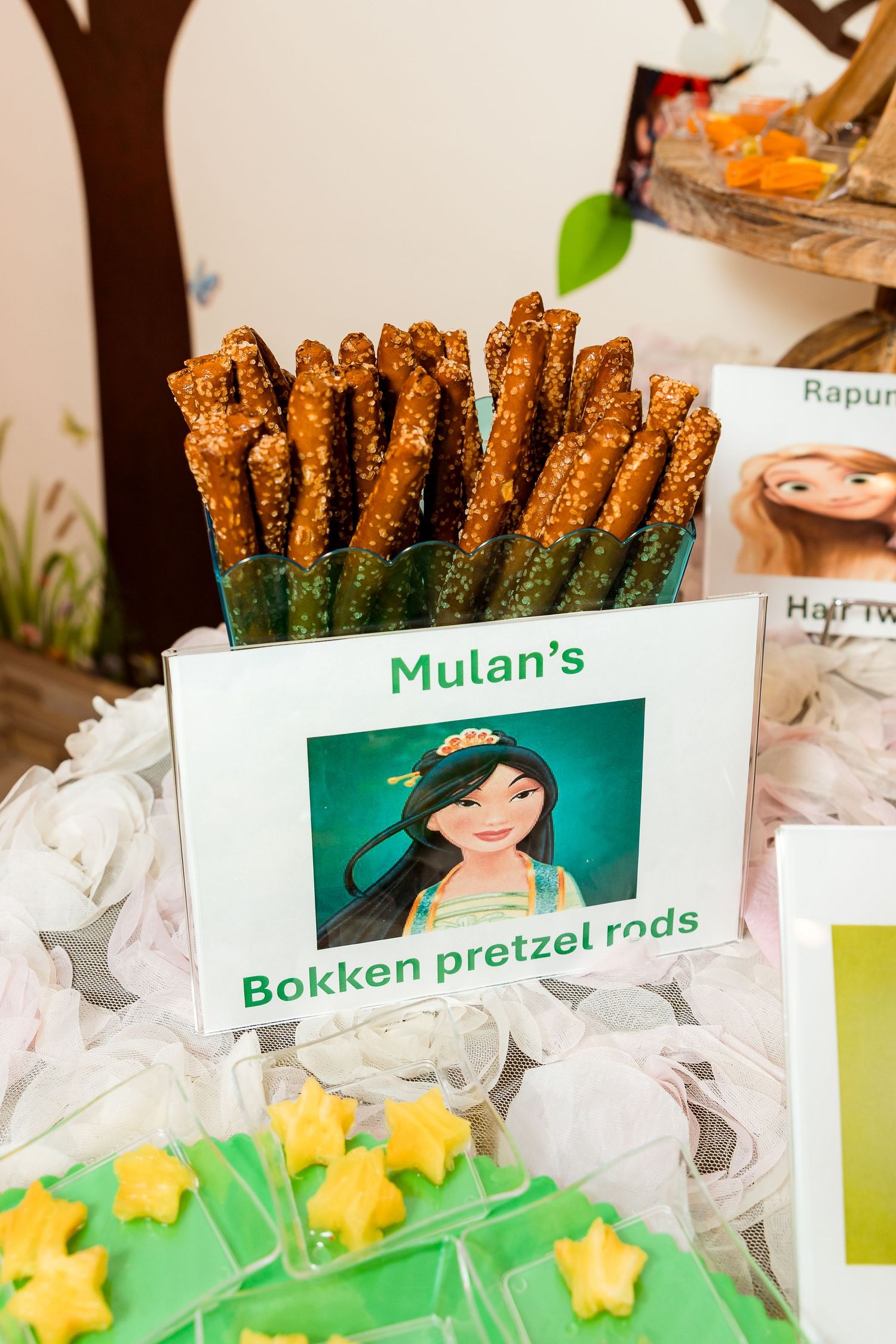 A themed party display featuring chocolate-covered pretzel rods labeled as Mulan's Bokken pretzel rods next to yellow star decorations.