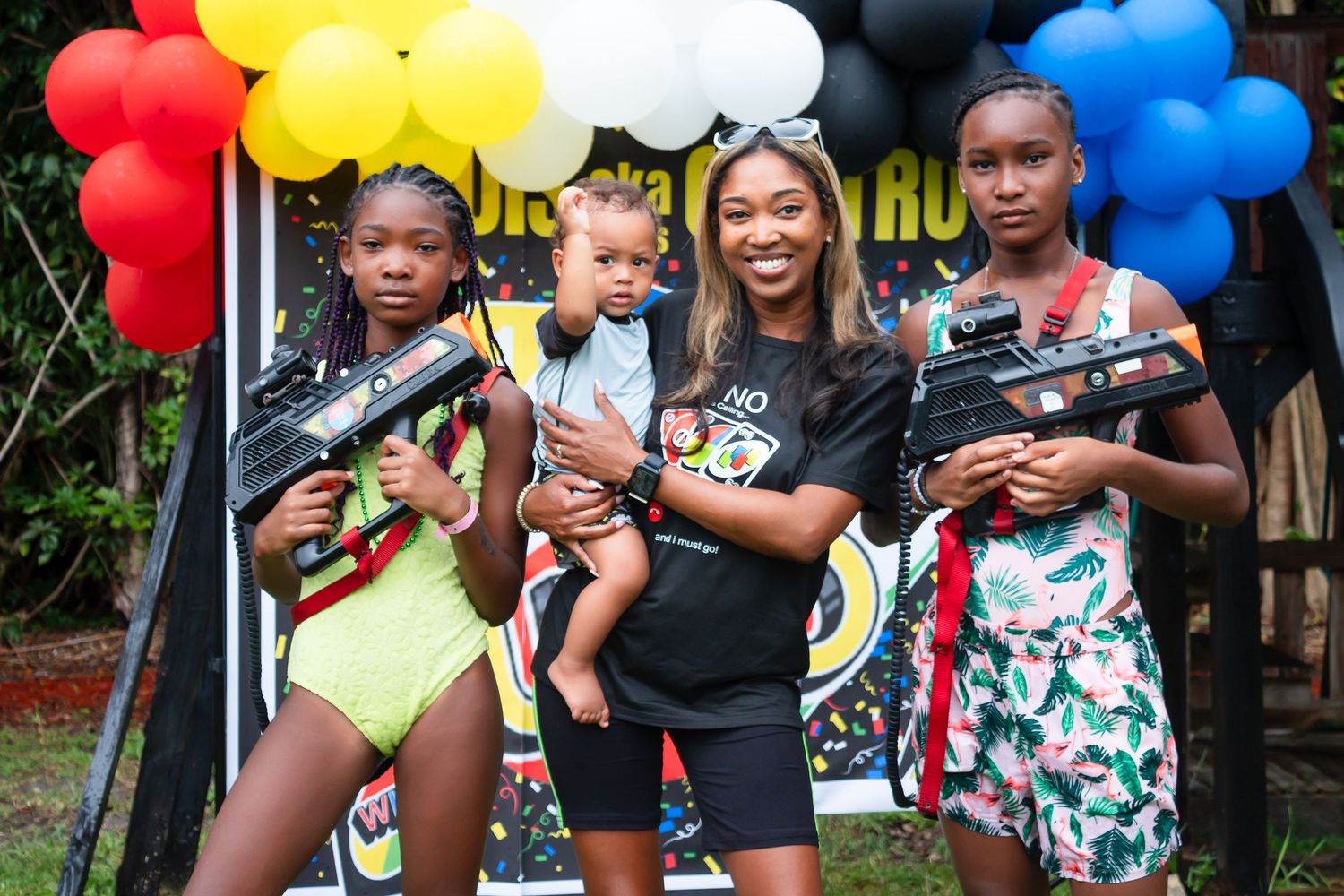 Group poses with colorful water guns at outdoor party with red, yellow, white and blue balloon decorations.