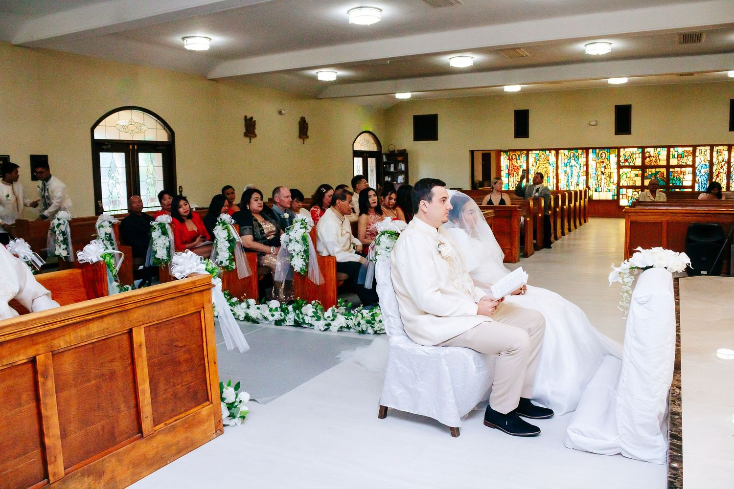Wedding ceremony setting with people in white attire seated in church pews.