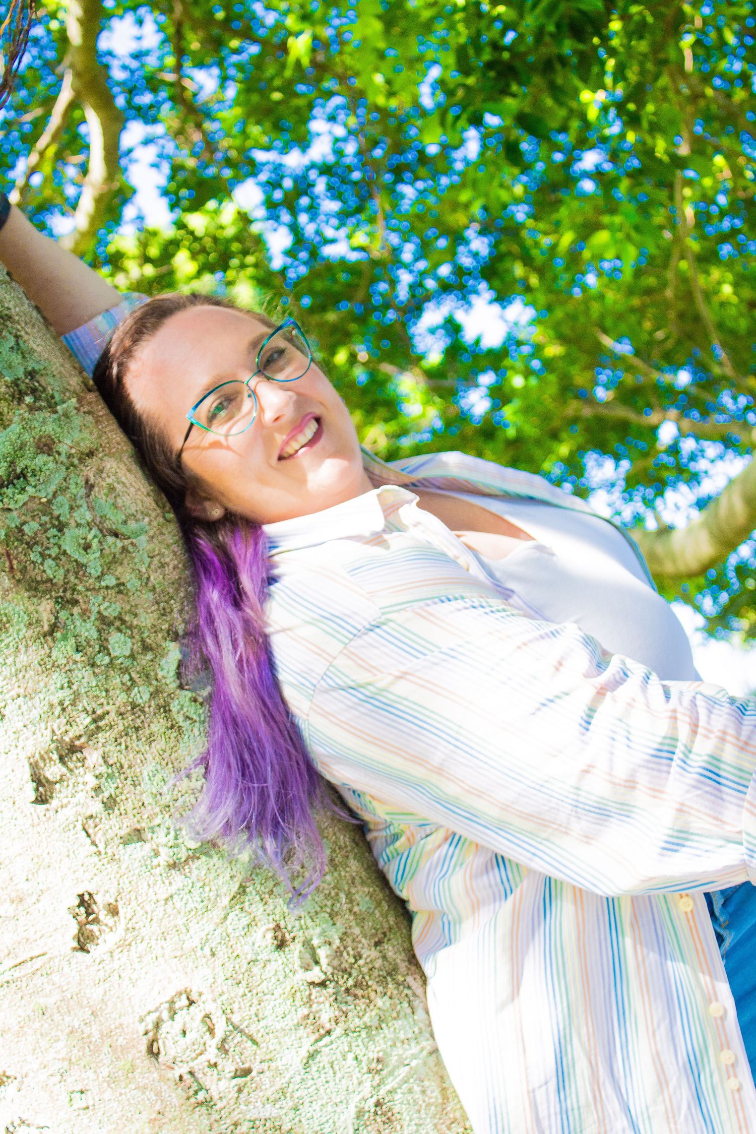 Artist in striped shirt leaning against tree trunk with sunny garden backdrop.