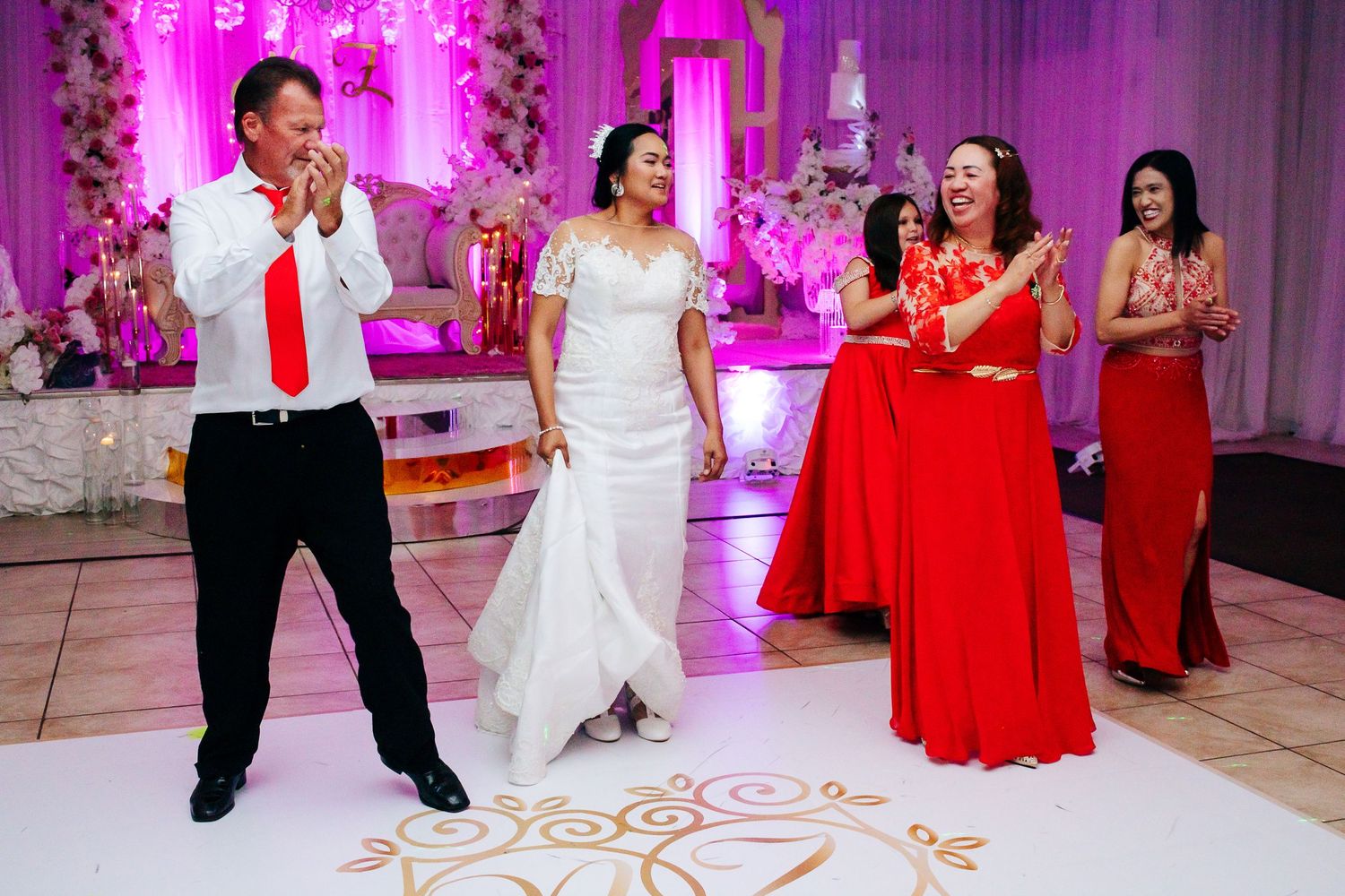 Wedding celebration with dancing guests in formal attire including red dresses against pink-lit decor.