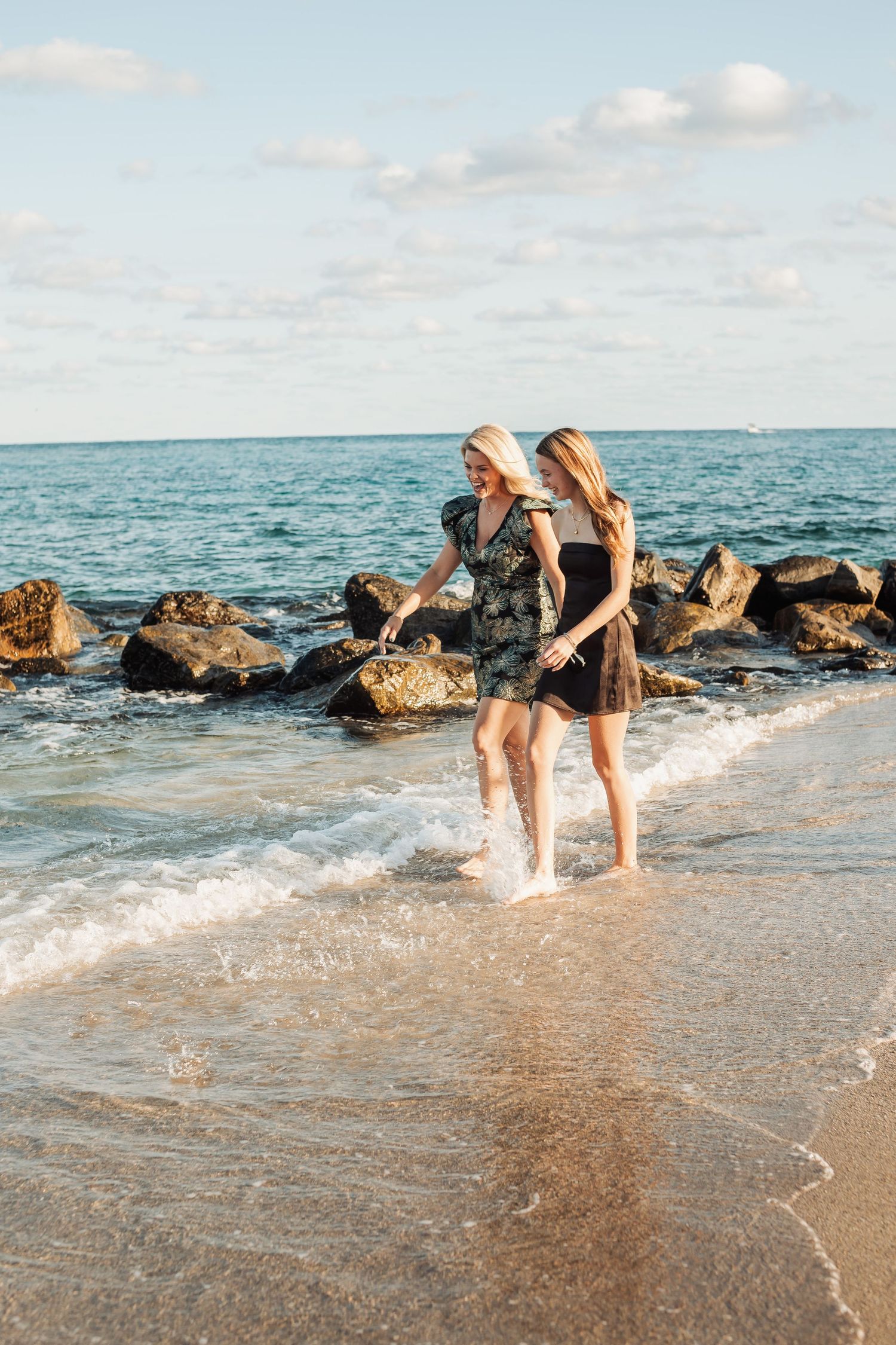 Two people in summer dresses splashing in ocean waves near rocky shore during sunset.