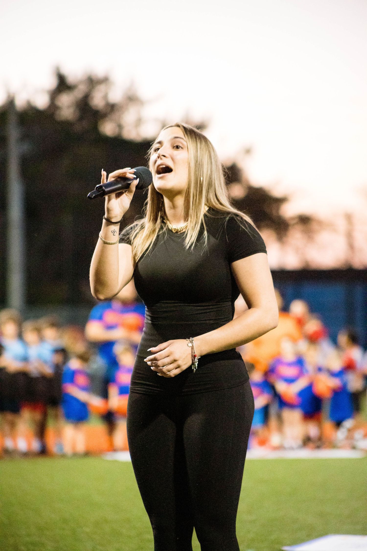 Performer sings national anthem on sports field at dusk wearing all black outfit.