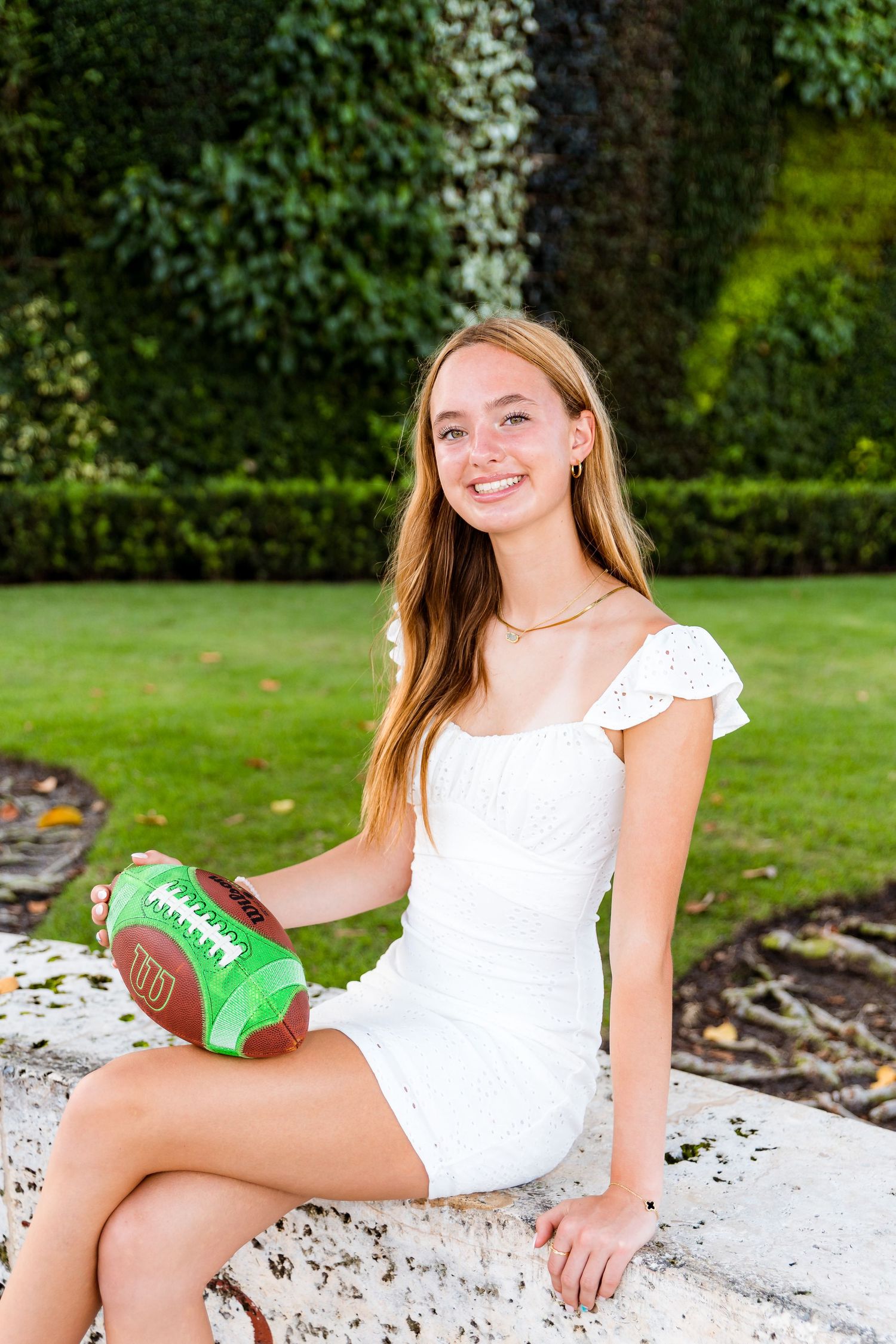A football is held by someone in a white eyelet lace dress sitting on a stone ledge in a garden setting.