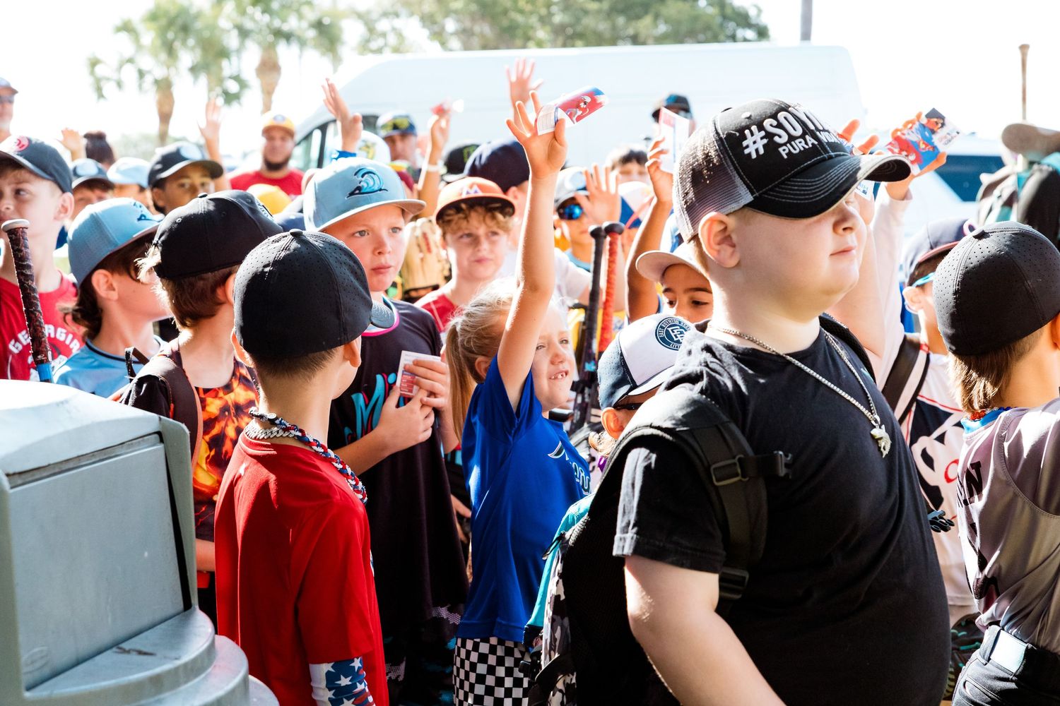 Excited crowd of fans raise their hands at an outdoor event with a security guard in a black shirt standing watch.