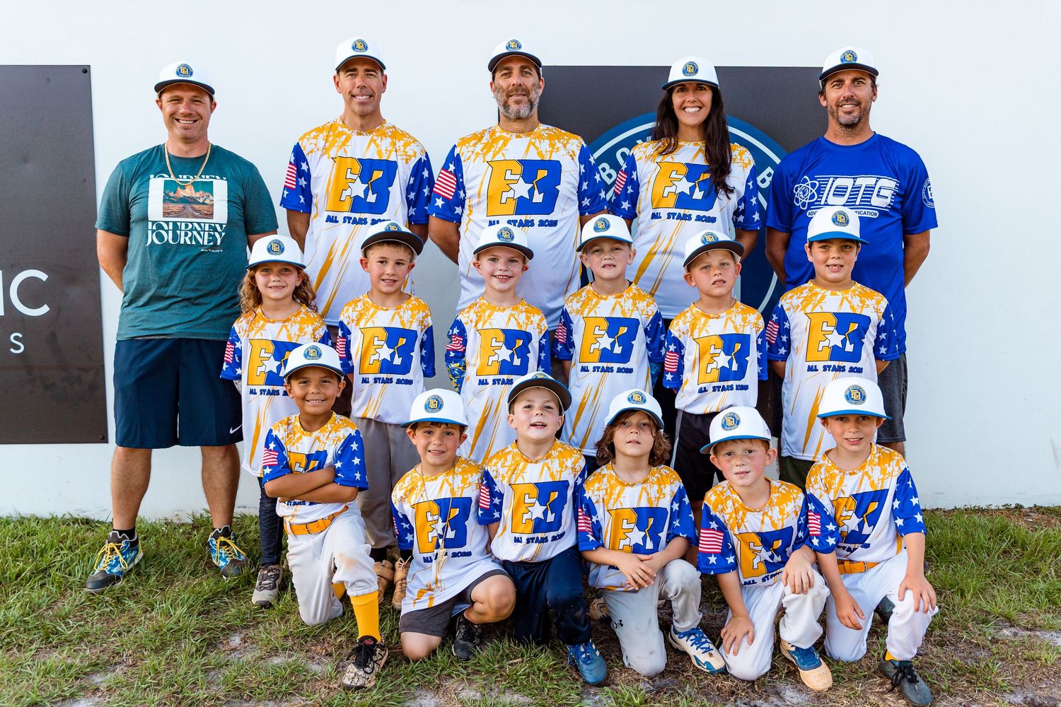 Youth baseball team in blue and yellow uniforms poses with their coaches for a group photo by a wall outdoors.