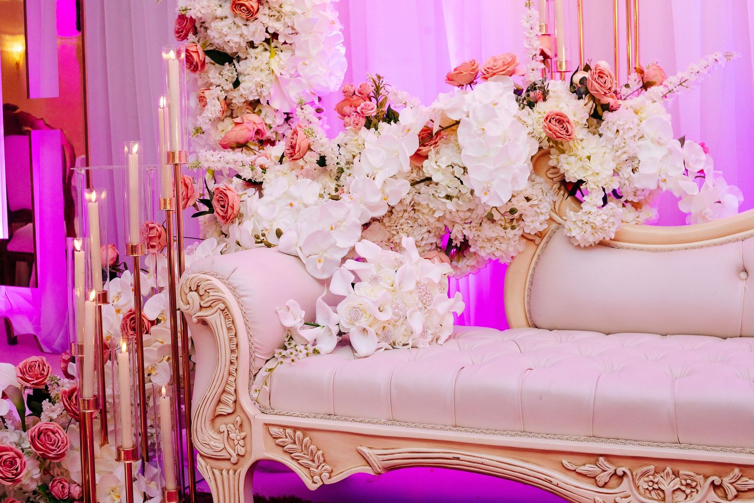 Luxurious white and gold wedding stage sofa decorated with pink and white floral arrangements and purple lighting.