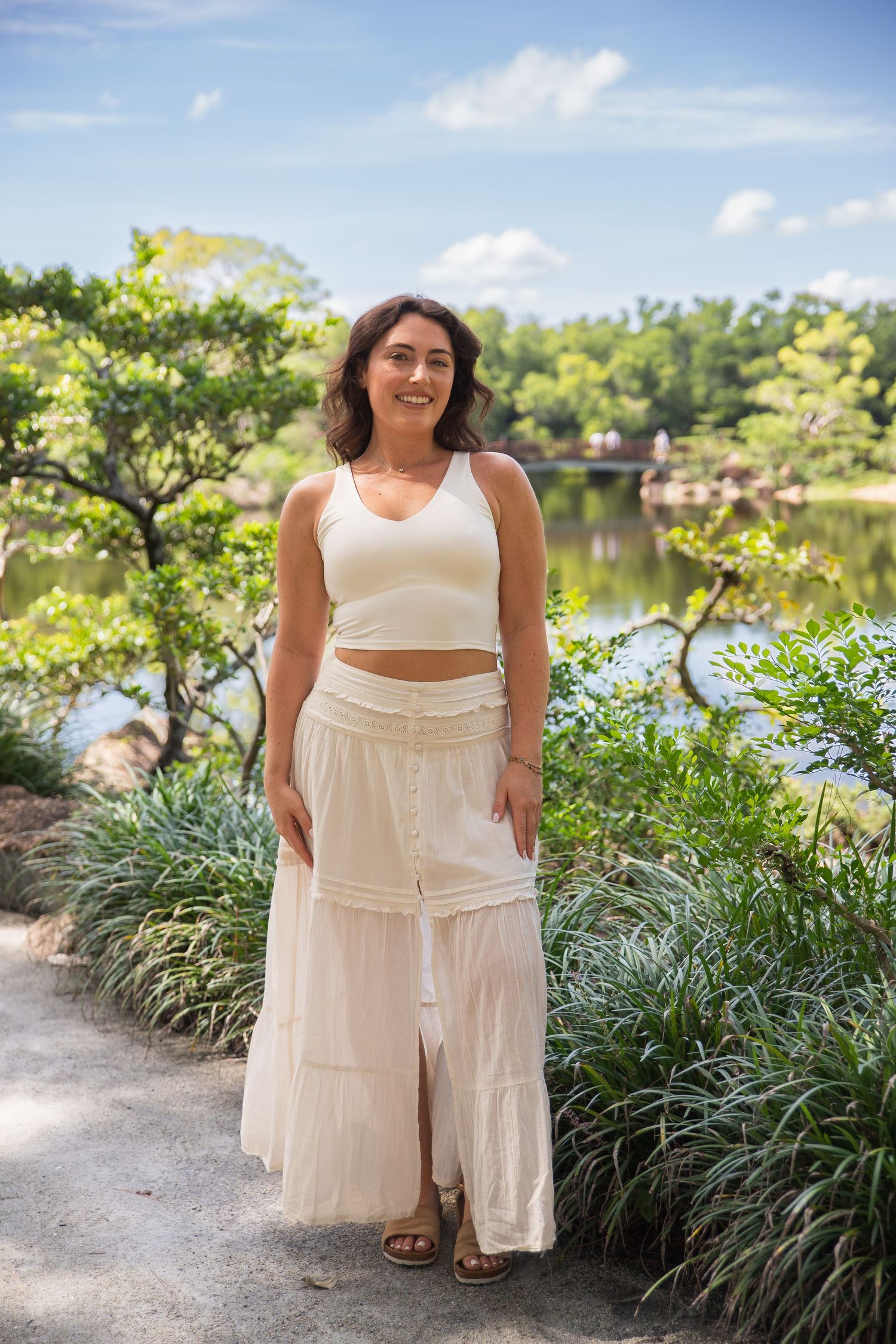 Woman wearing white outfit poses gracefully by lush greenery and water on sunny day.