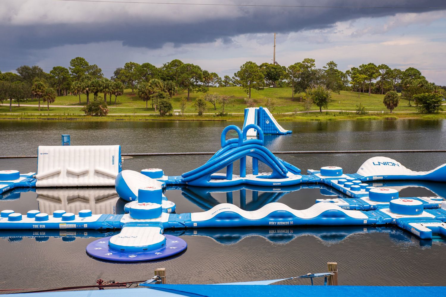 Large inflatable water park with blue and white obstacles floating on a lake surrounded by trees and green landscape.