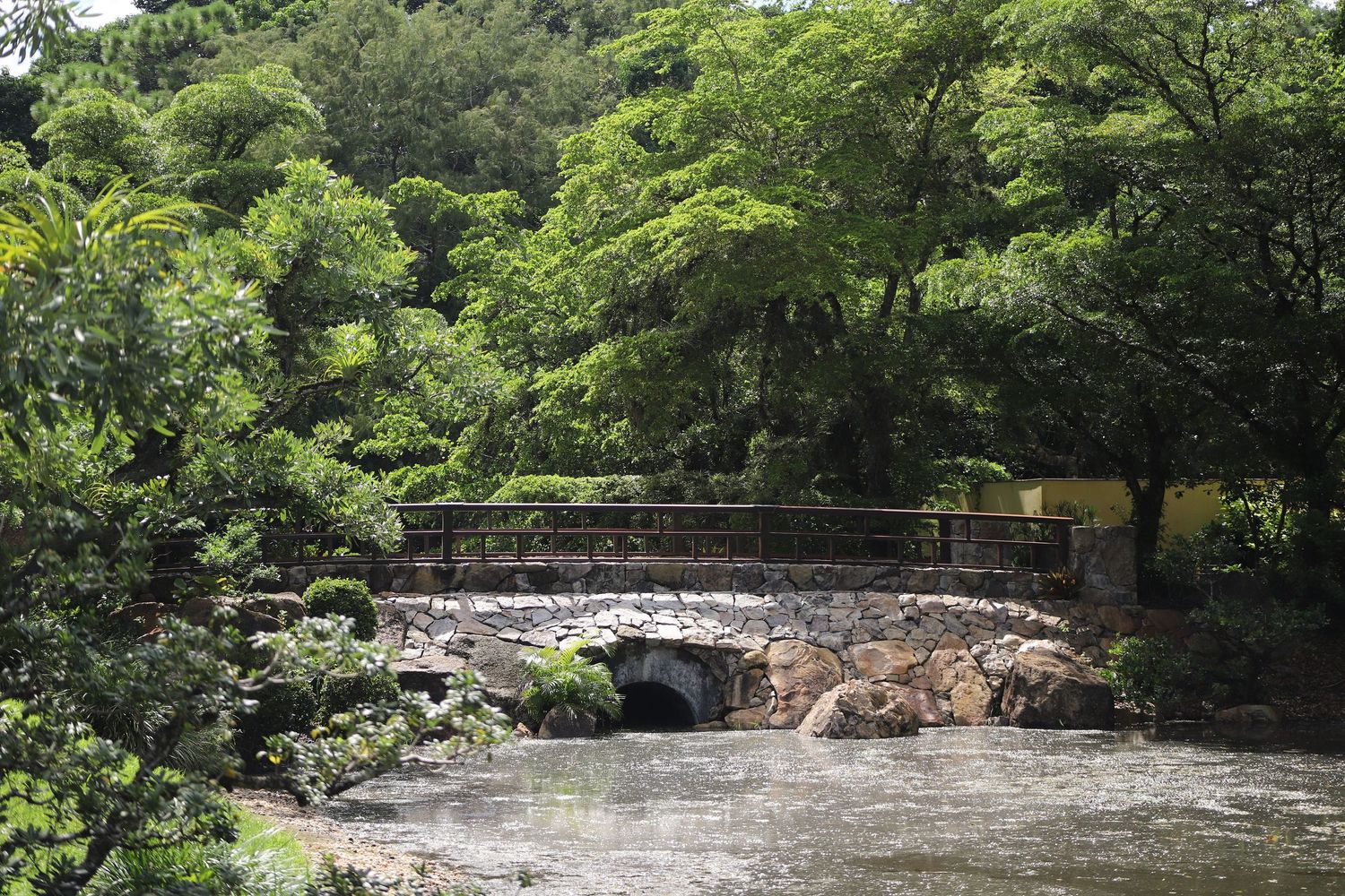 A scenic stone bridge spans over a flowing stream surrounded by lush green trees and foliage in a natural park setting.