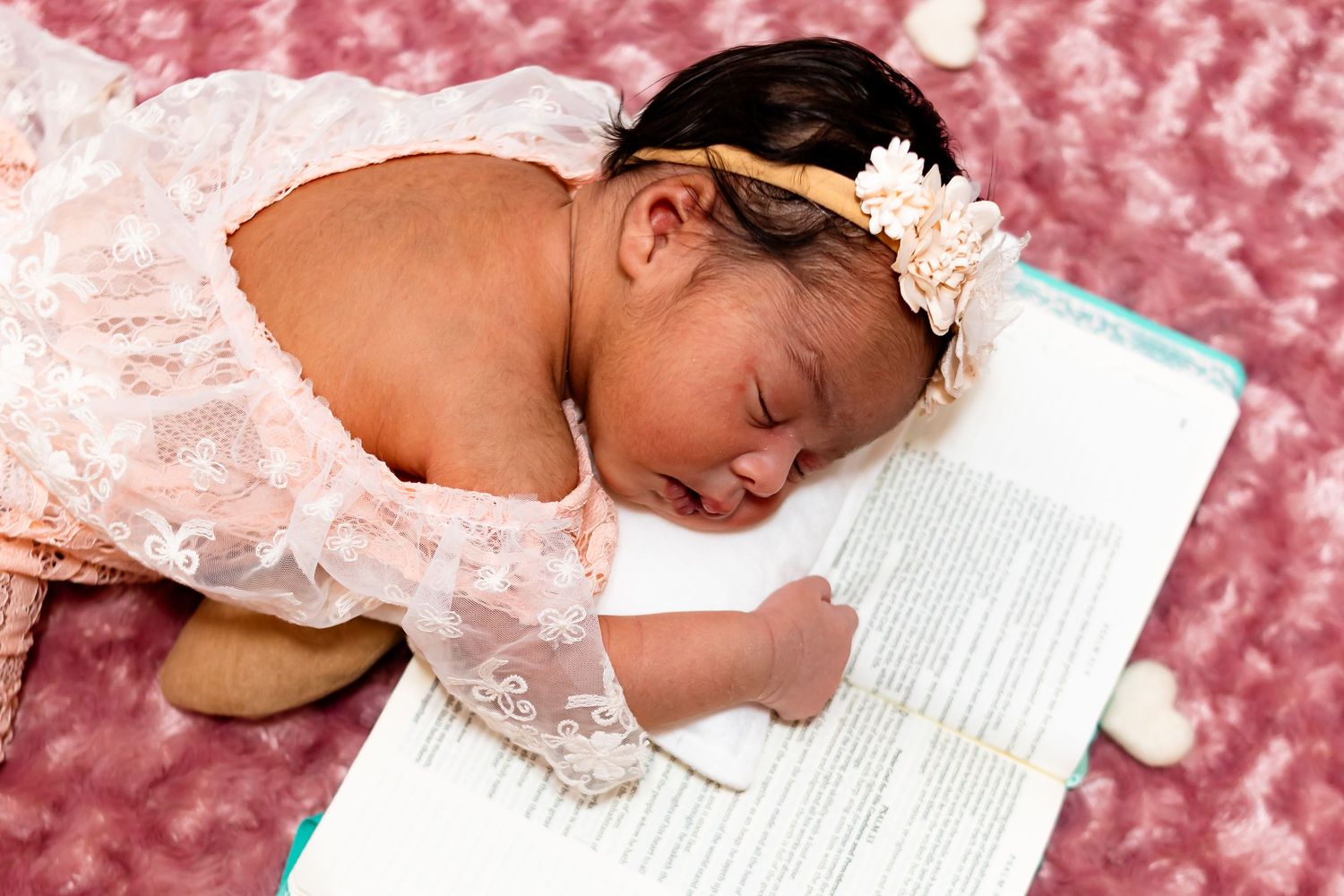 Newborn baby girl in pink lace dress with floral headband sleeping on an open book.