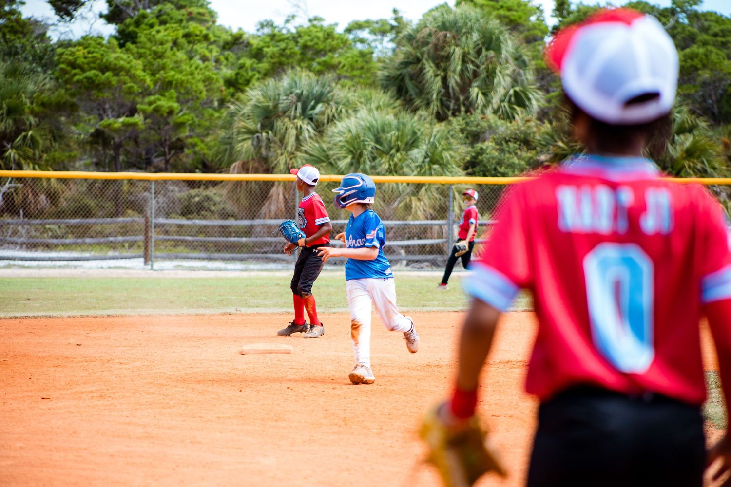 Baseball game action on dirt field with players in red and blue uniforms under sunny skies.