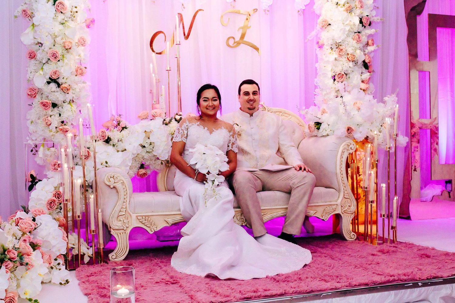 Elegant wedding couple sits on ornate sofa with pink lighting and floral arch decorations at dreamy reception setup.