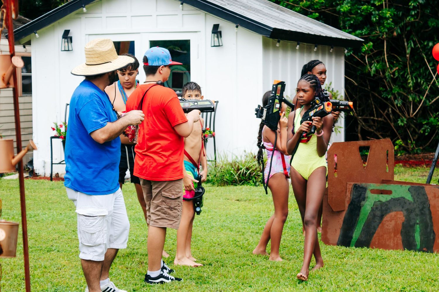Party guests playing outdoor games in a backyard setting with white building in background.