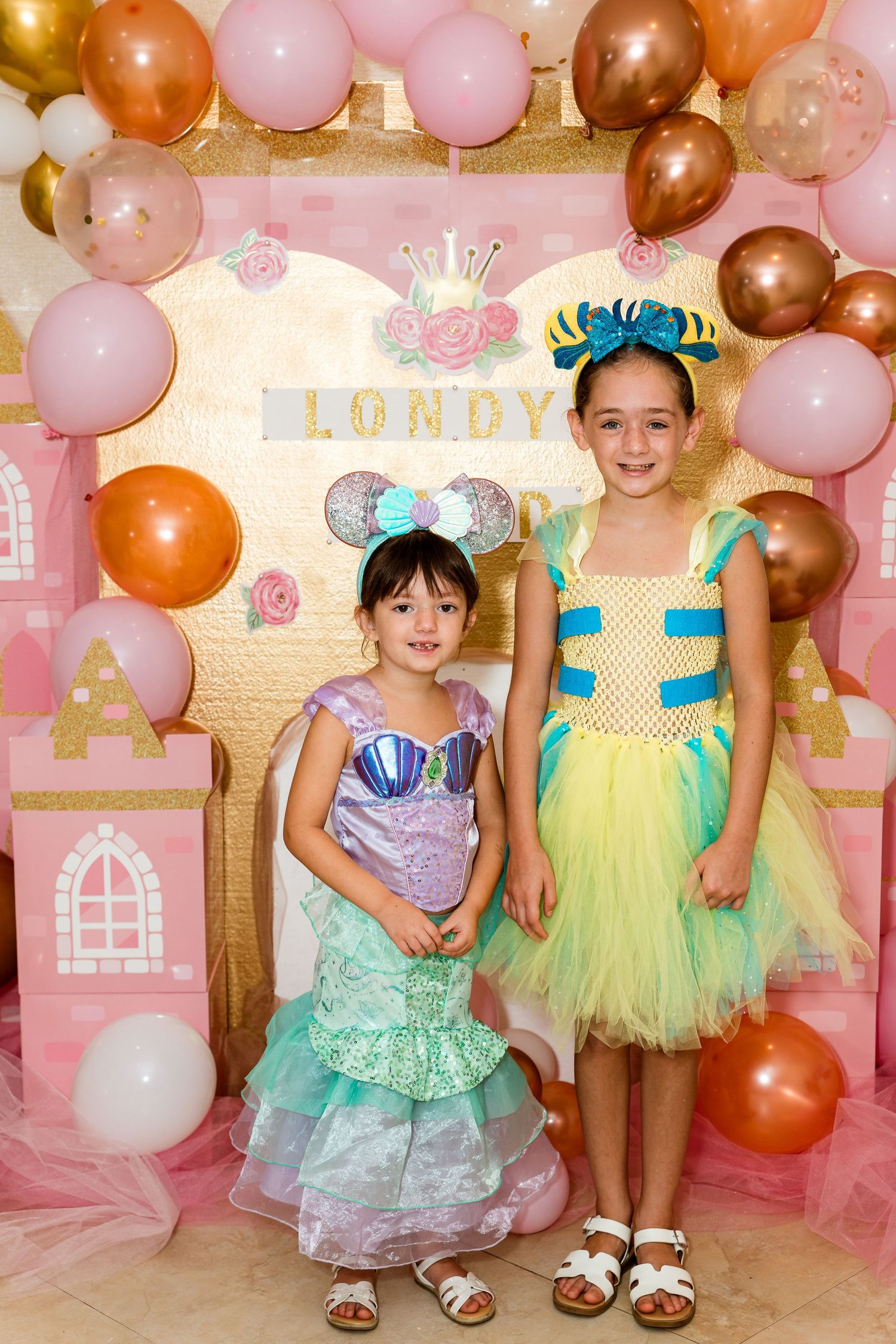 Two young guests in fancy princess dresses pose in front of a pink balloon arch and castle backdrop at a party.