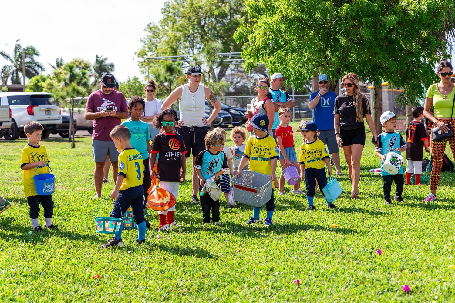 Children participate in an Easter egg hunt on a sunny green lawn with parents watching in the background.