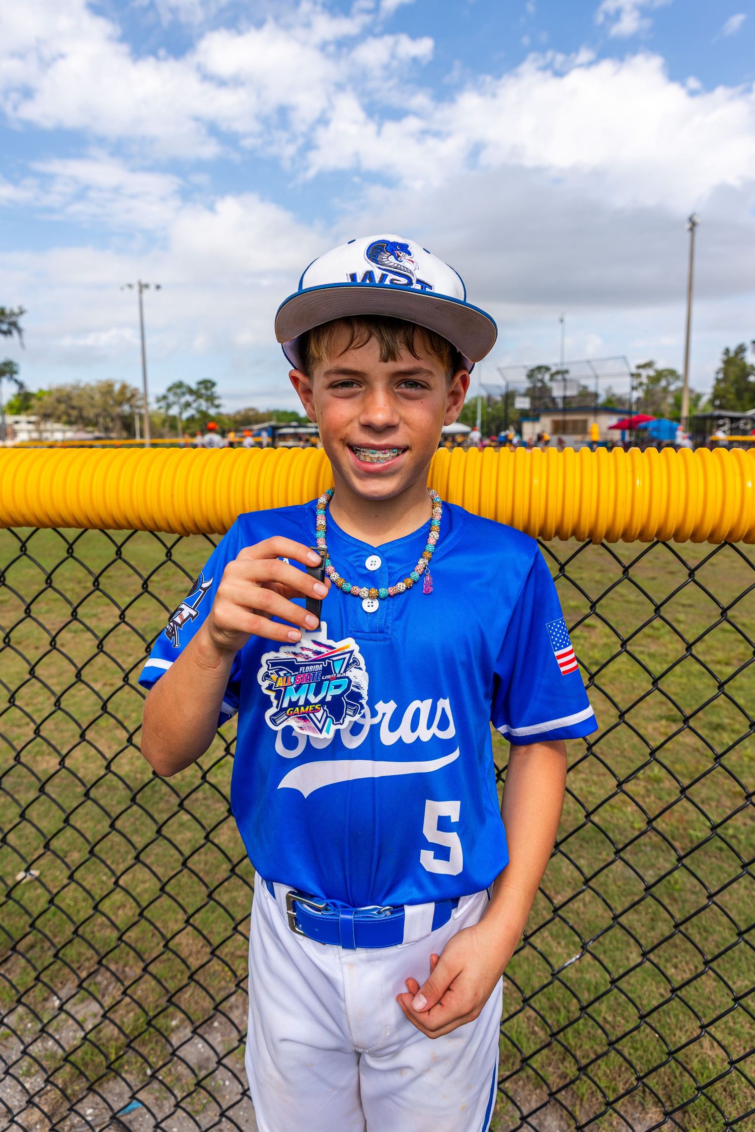 Young baseball player in blue and white uniform poses in front of chain link fence on sunny day.