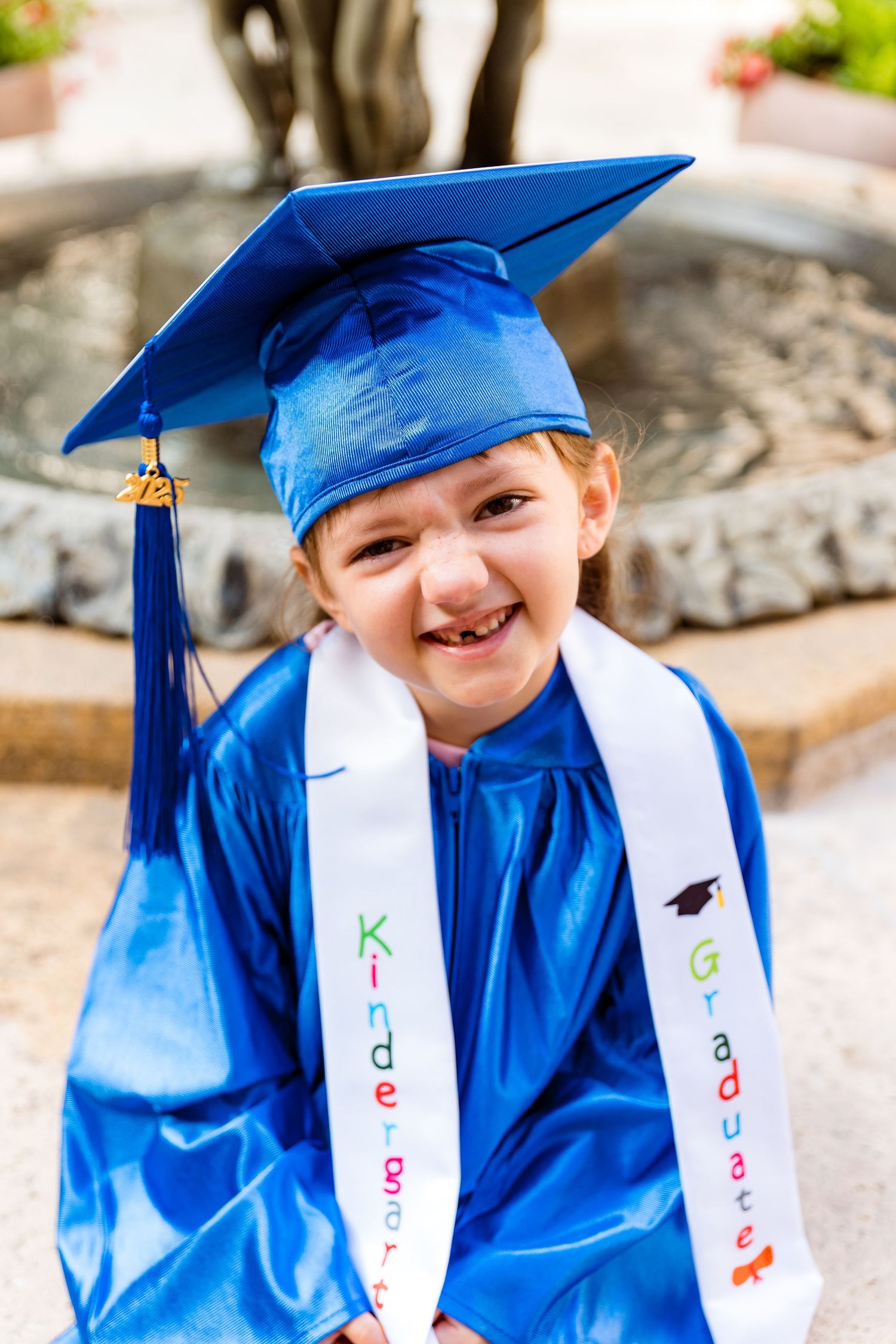 A young graduate in shiny blue cap and gown with decorated stoles smiles while sitting outdoors by a fountain.