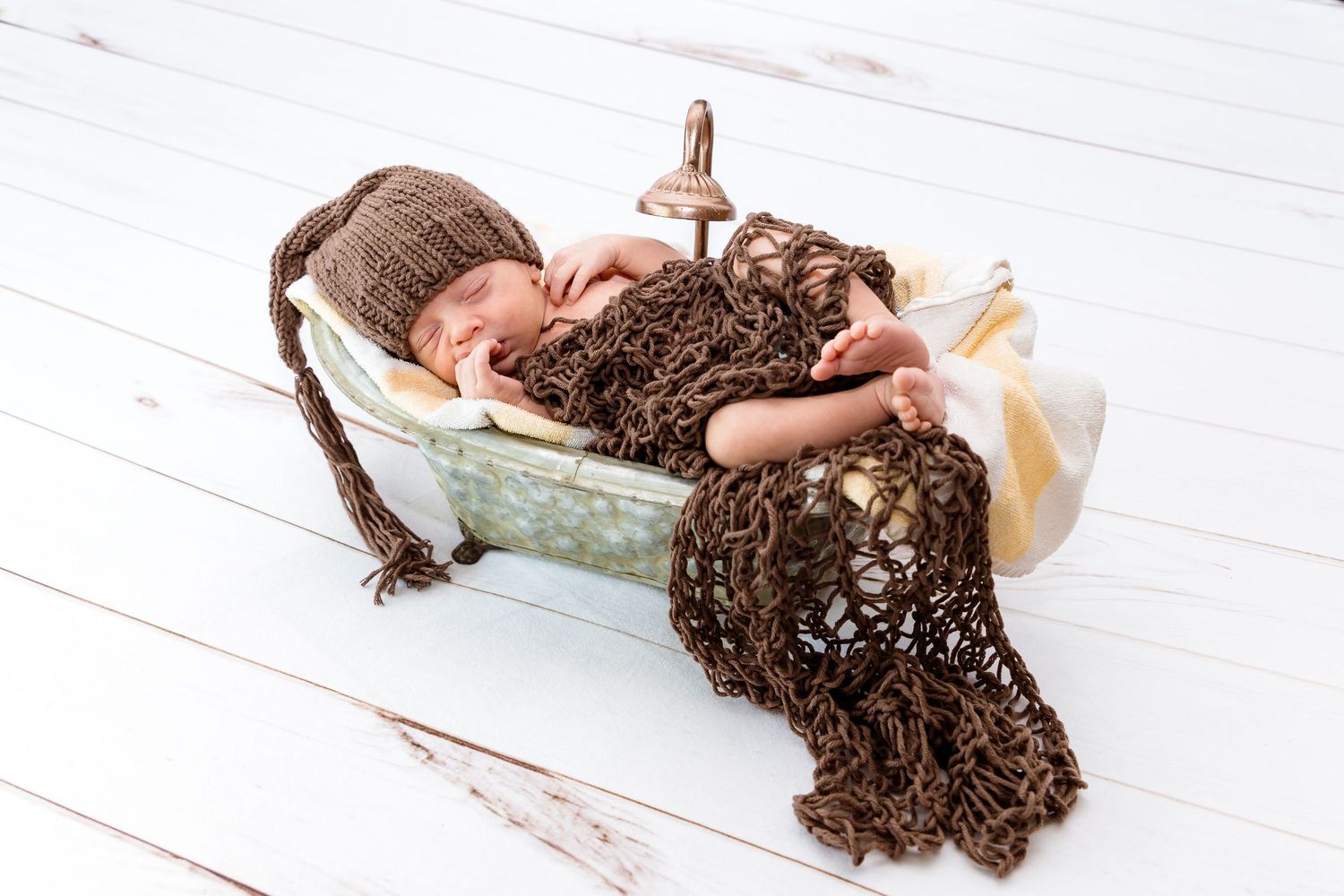 A newborn rests on a small mint-colored bed while covered in a brown lace-textured fabric on white wooden flooring.