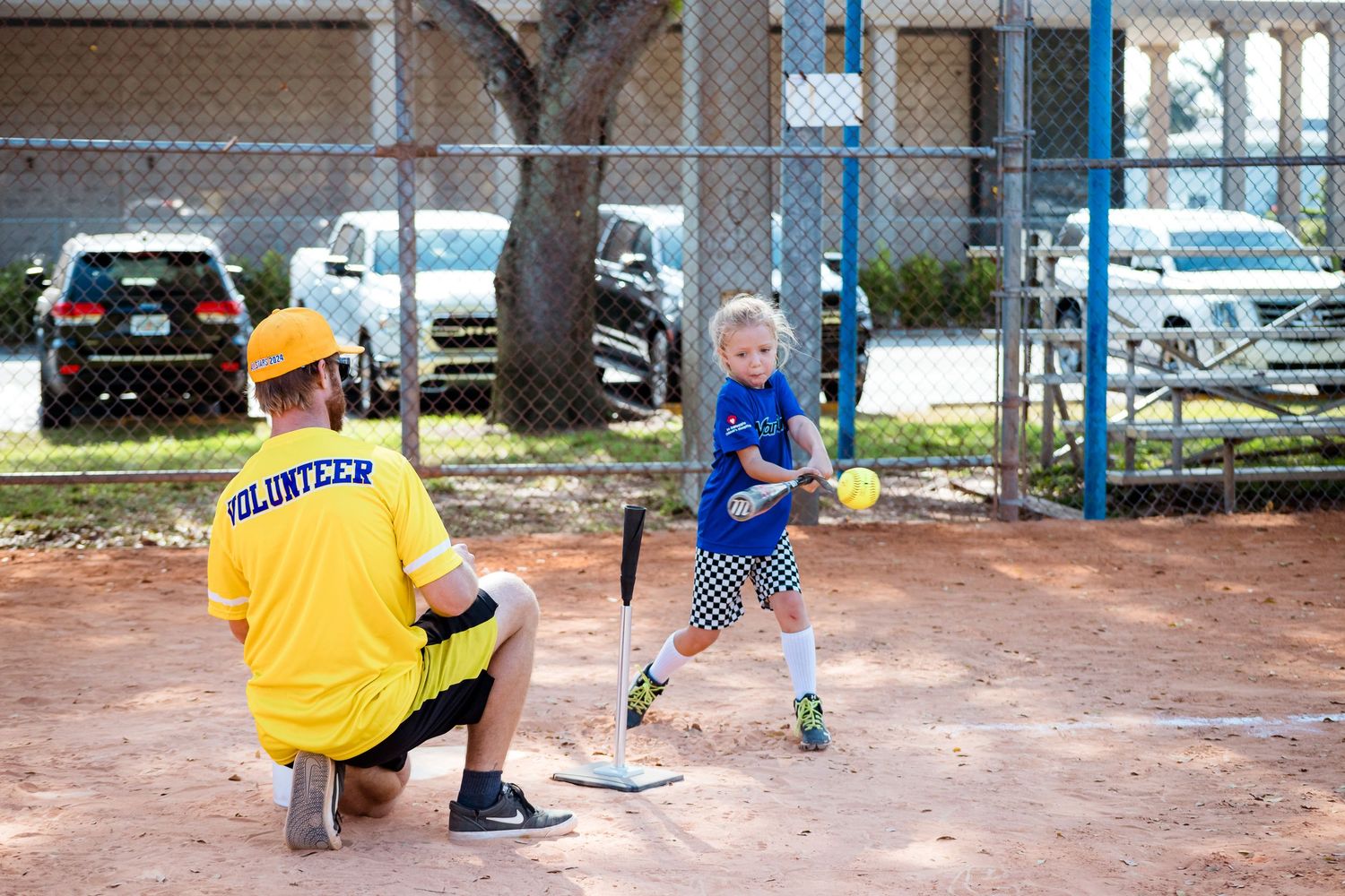 A youth baseball player practices batting with a coach crouched nearby at a dirt field with picnic tables and cars in background.