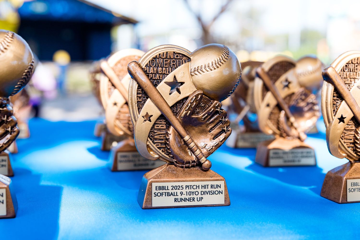 Row of gold baseball and softball sports trophies with crossed bats design displayed on bright blue table surface.